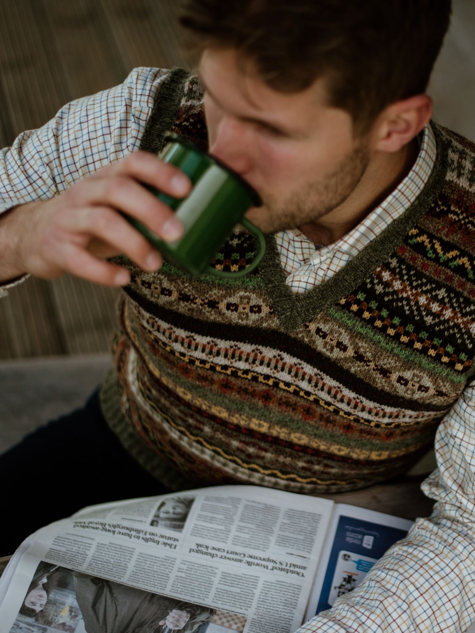 A man in a Campbells of Beauly Fairisle Slipover and plaid shirt sits and drinks from a green mug while reading a newspaper. Shot from above, the image focuses on his sleeveless layer, cup, and paper with softly blurred details.