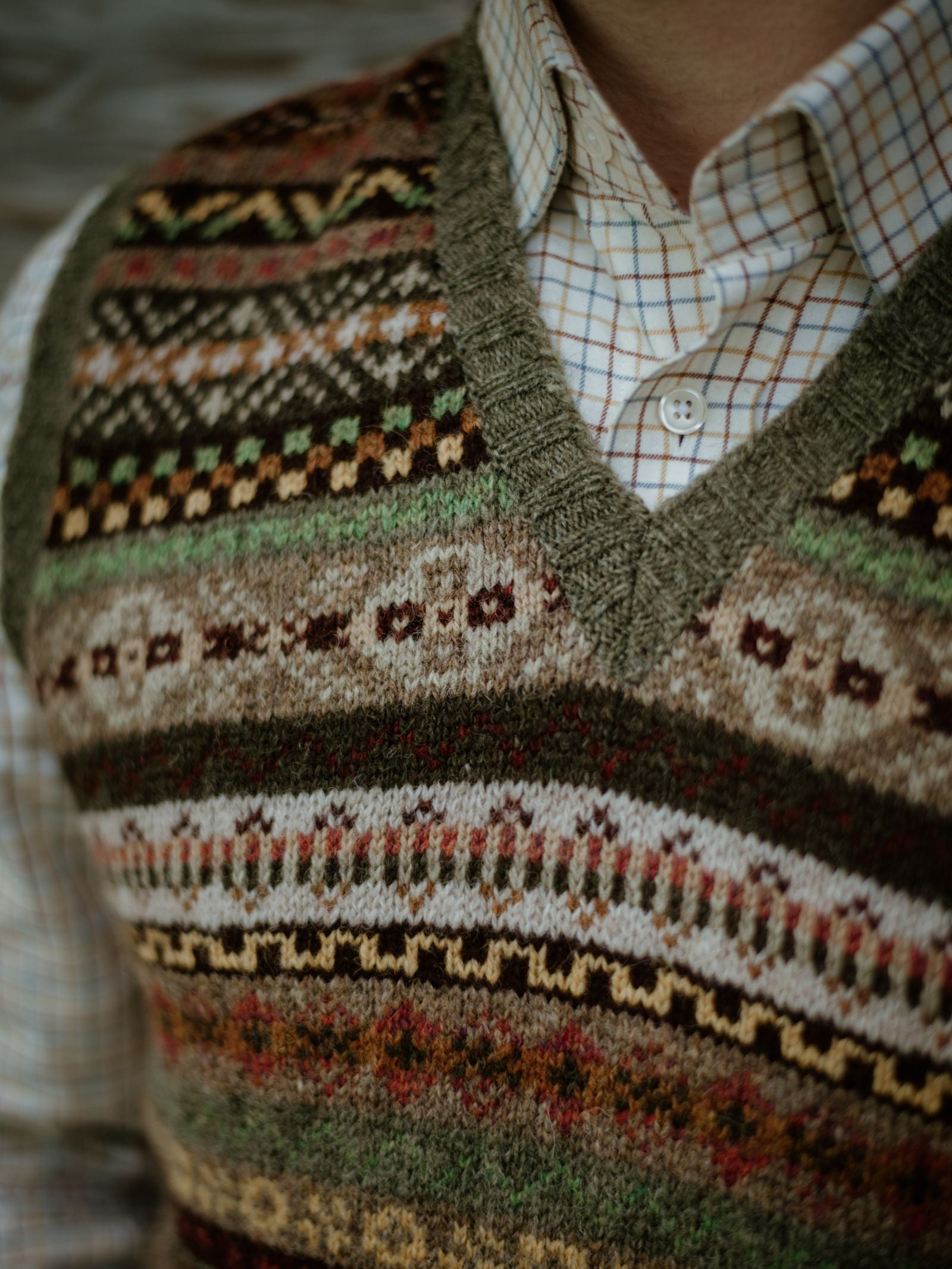 A person wears the Campbells of Beauly Fairisle Slipover—a colorful, sleeveless Shetland wool knit featuring green trim and intricate earthy-toned Fairisle patterns—over a plaid shirt; only the torso and part of the neck are visible.