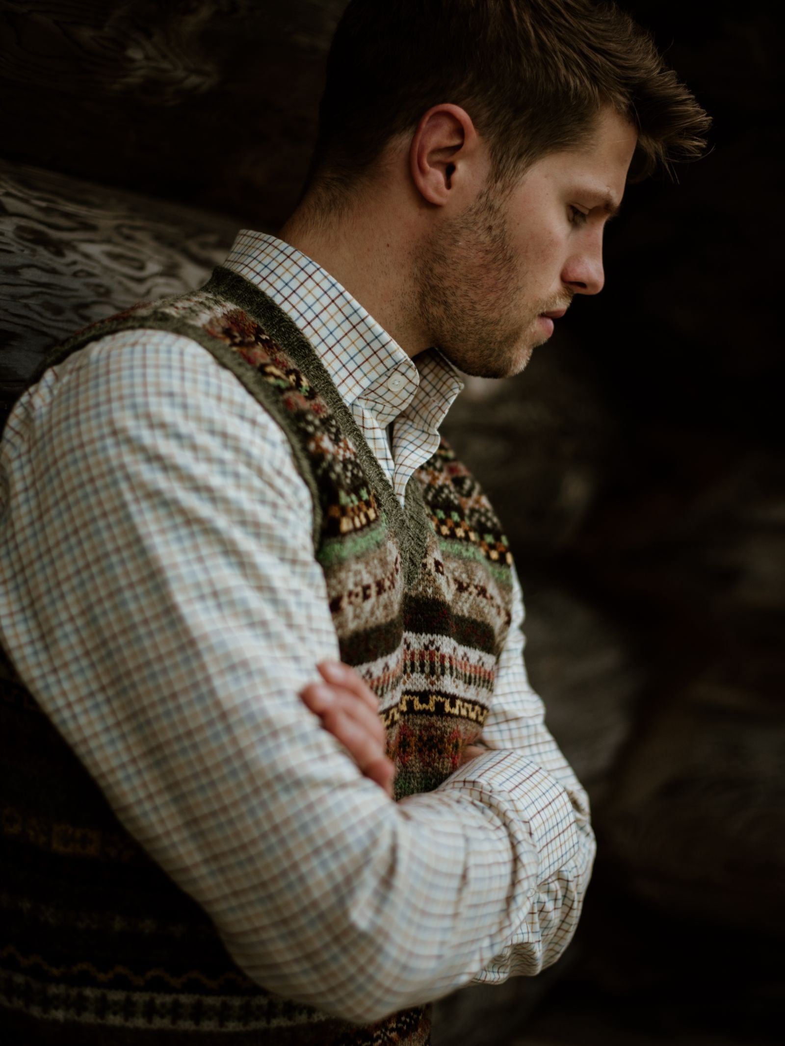 A thoughtful man in profile, arms crossed, wears a plaid shirt under a Campbells of Beauly Fairisle Slipover, set against a dark rustic wooden backdrop.