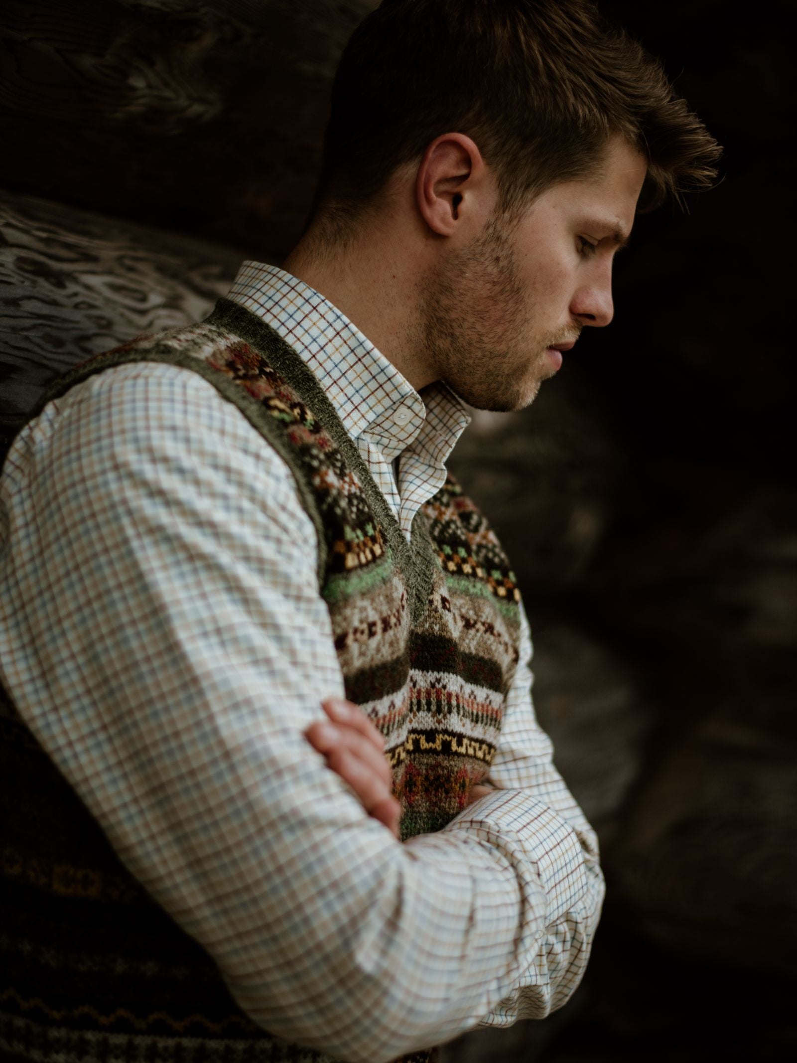 A young man with short brown hair stands in profile, arms crossed, wearing a Campbells of Beauly Windowpane Shirt under a patterned sweater vest. He gazes down thoughtfully against a dark wooden background, softly lit for a contemplative mood.
