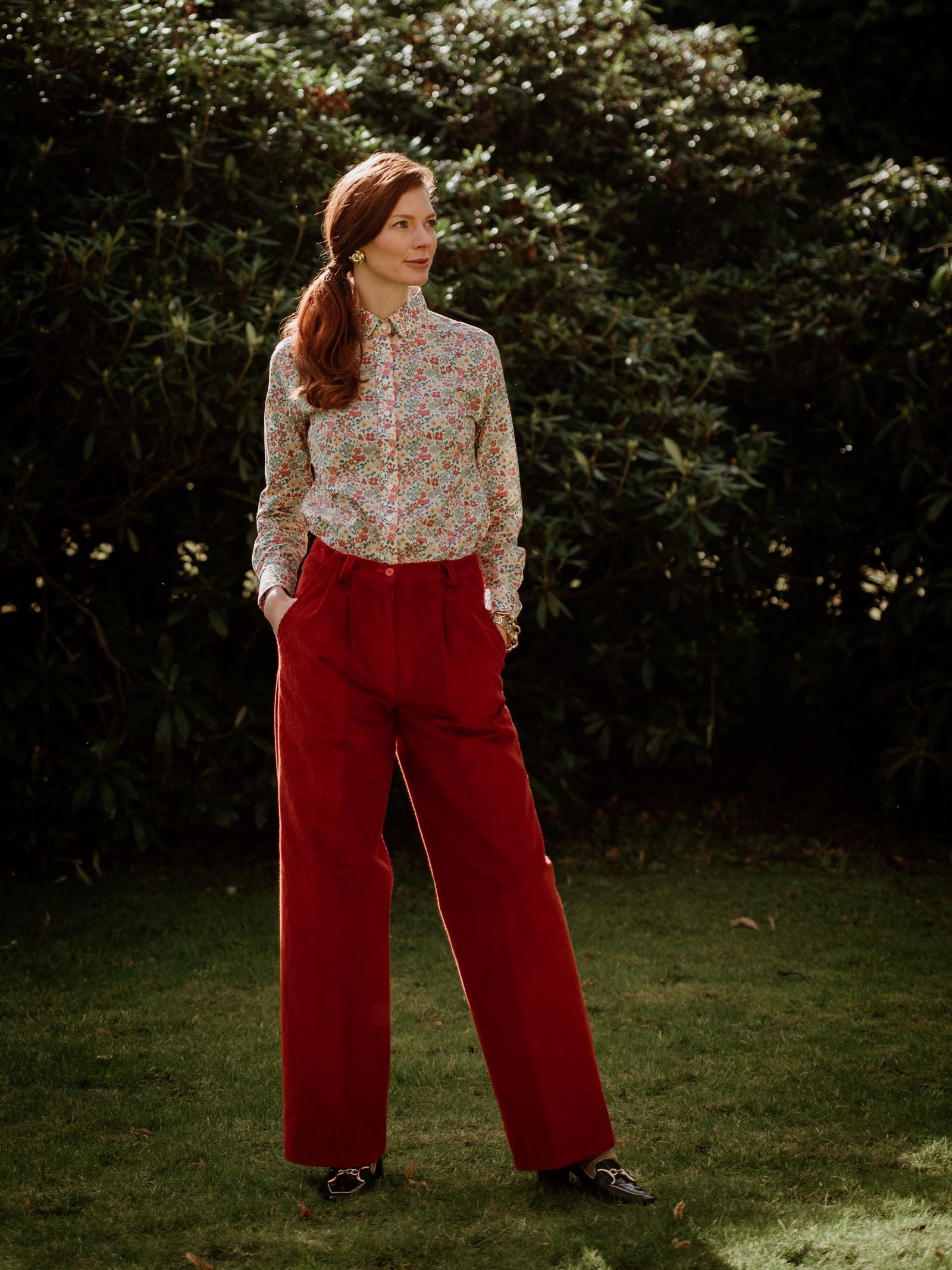 A woman stands on grass before lush greenery, wearing a long-sleeve floral blouse tucked into Campbells of Beauly Wide Leg Cords. Her long auburn hair is pulled back as she gazes thoughtfully to her right.