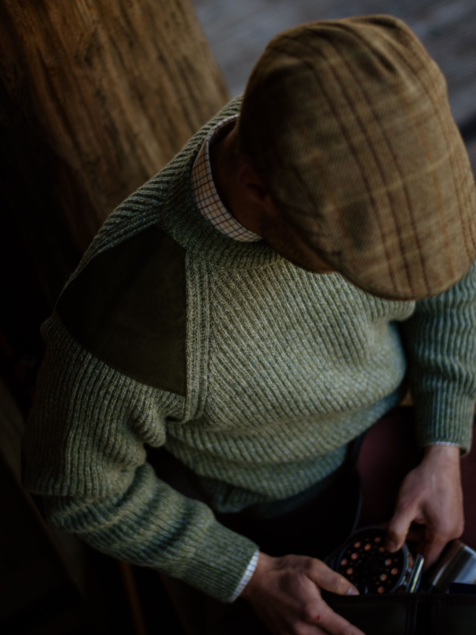 Wearing the Campbells of Beauly Scotia Suede Jumper, paired with a plaid shirt and light brown plaid flat cap, someone gazes down at a small container against a rustic blurred wood background, their face not visible.