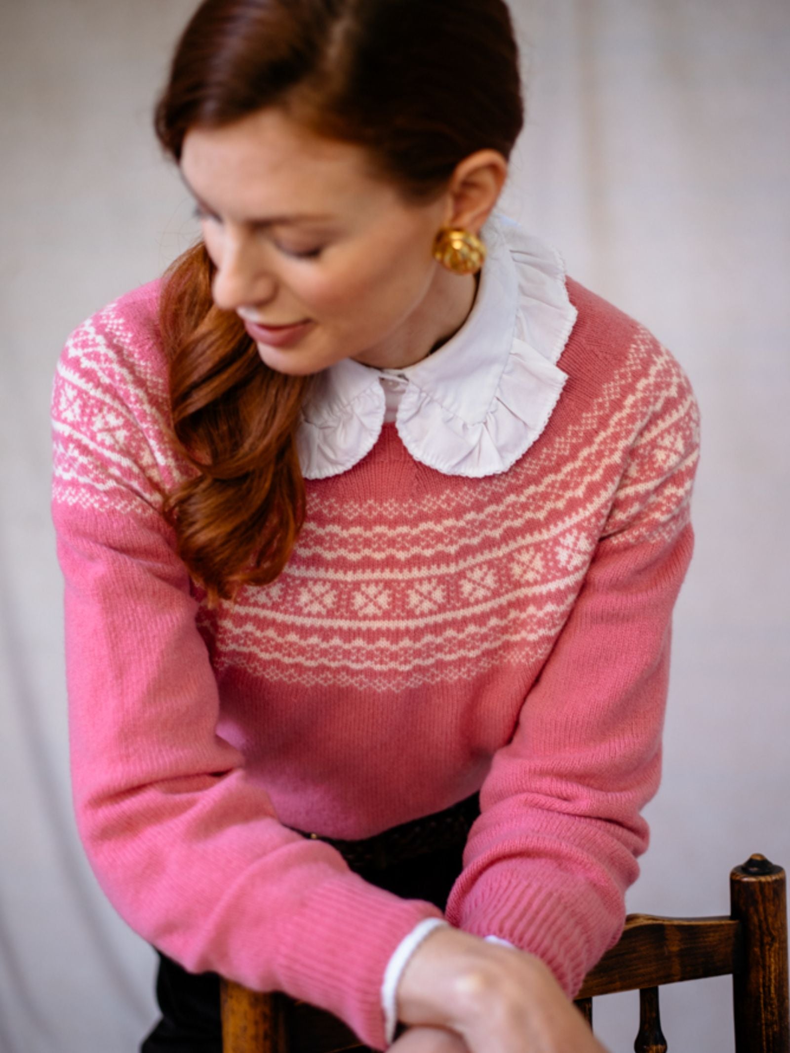 A woman with long auburn hair wears the Campbells of Beauly Two-Colour Fairisle Yoke Jumper in pink superfine Geelong lambswool over a white blouse, paired with gold round earrings. She sits on a wooden chair, looking down against a neutral background.
