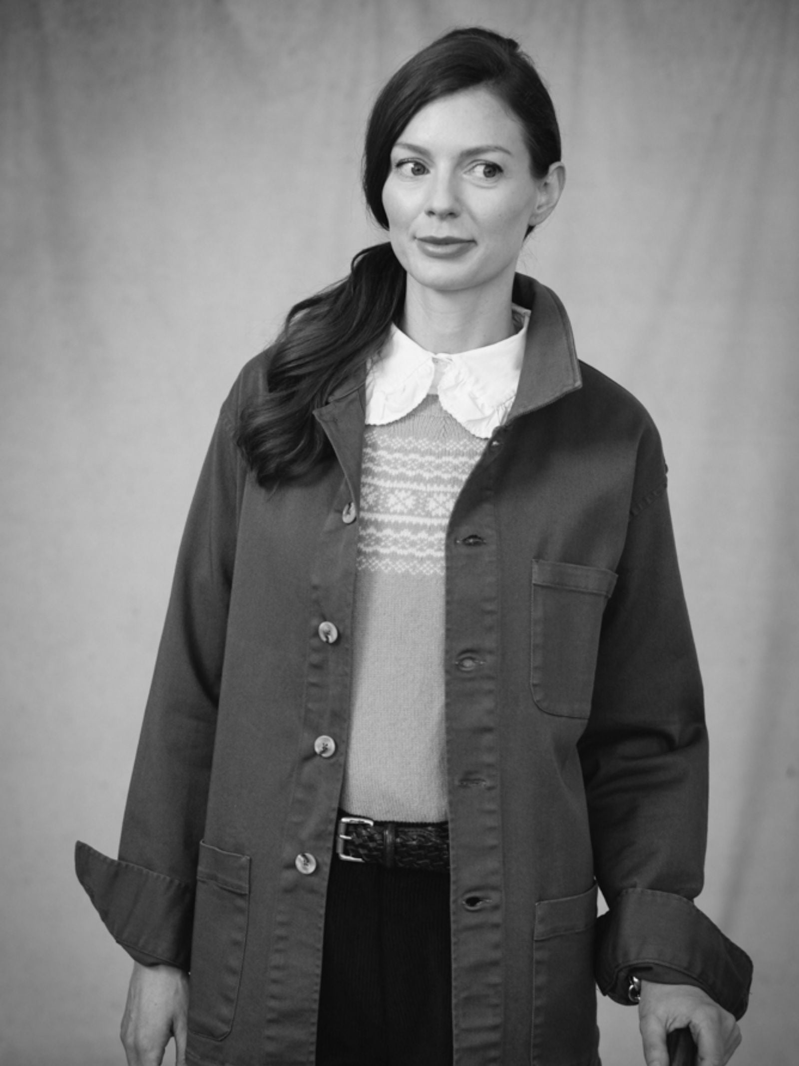 Black and white photo of a woman in a relaxed fit Summer Chore Jacket by Campbells of Beauly, layered over a collared shirt and sweater. With wavy hair styled to one side, she gazes thoughtfully to her right before a plain backdrop.