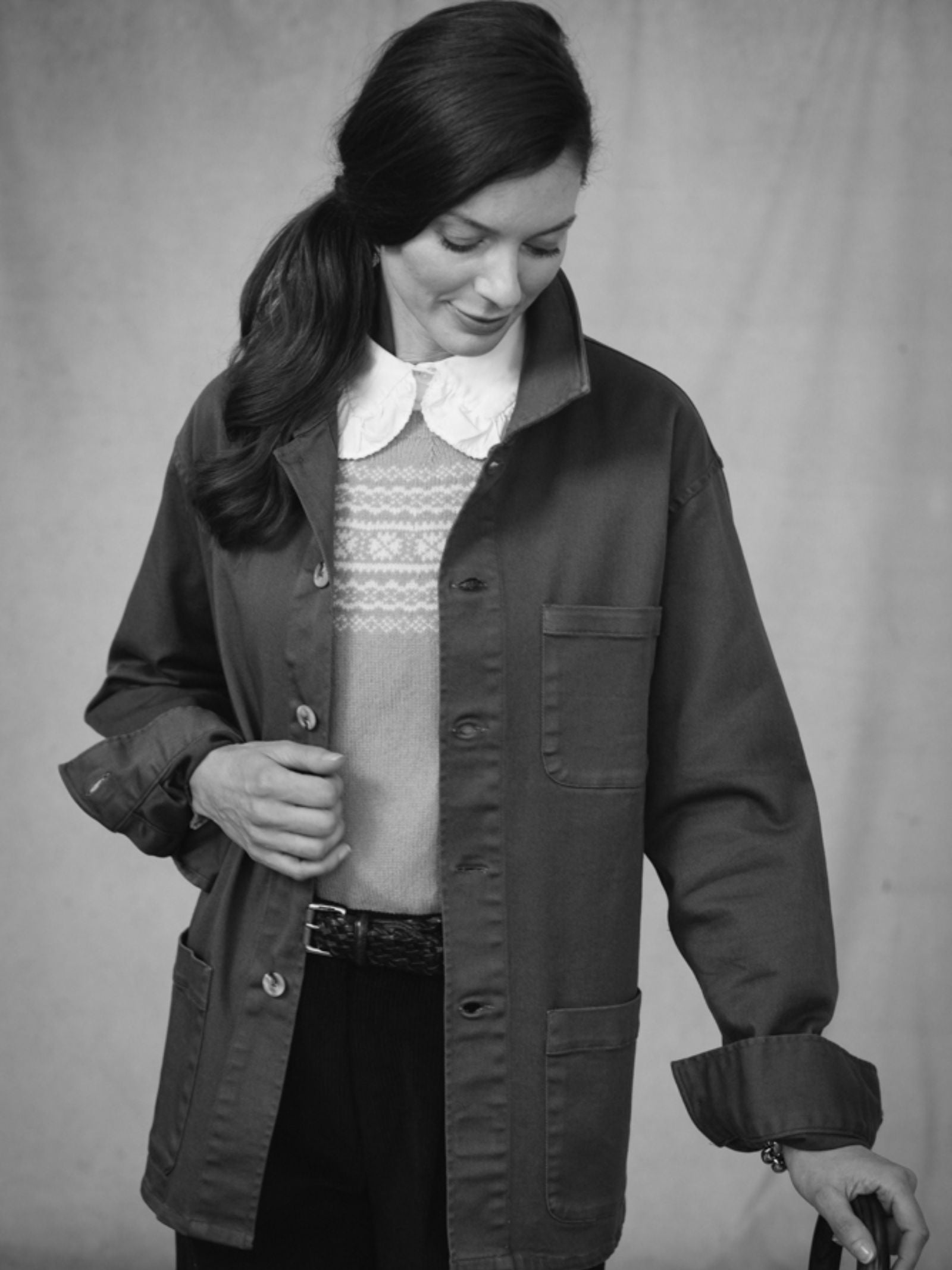 A woman with dark hair in a side ponytail smiles softly as she looks down, wearing a relaxed fit collared shirt under the Campbells of Beauly Summer Chore Jacket. Her left hand holds the lapel; her right arm is bent. The photo is black and white.