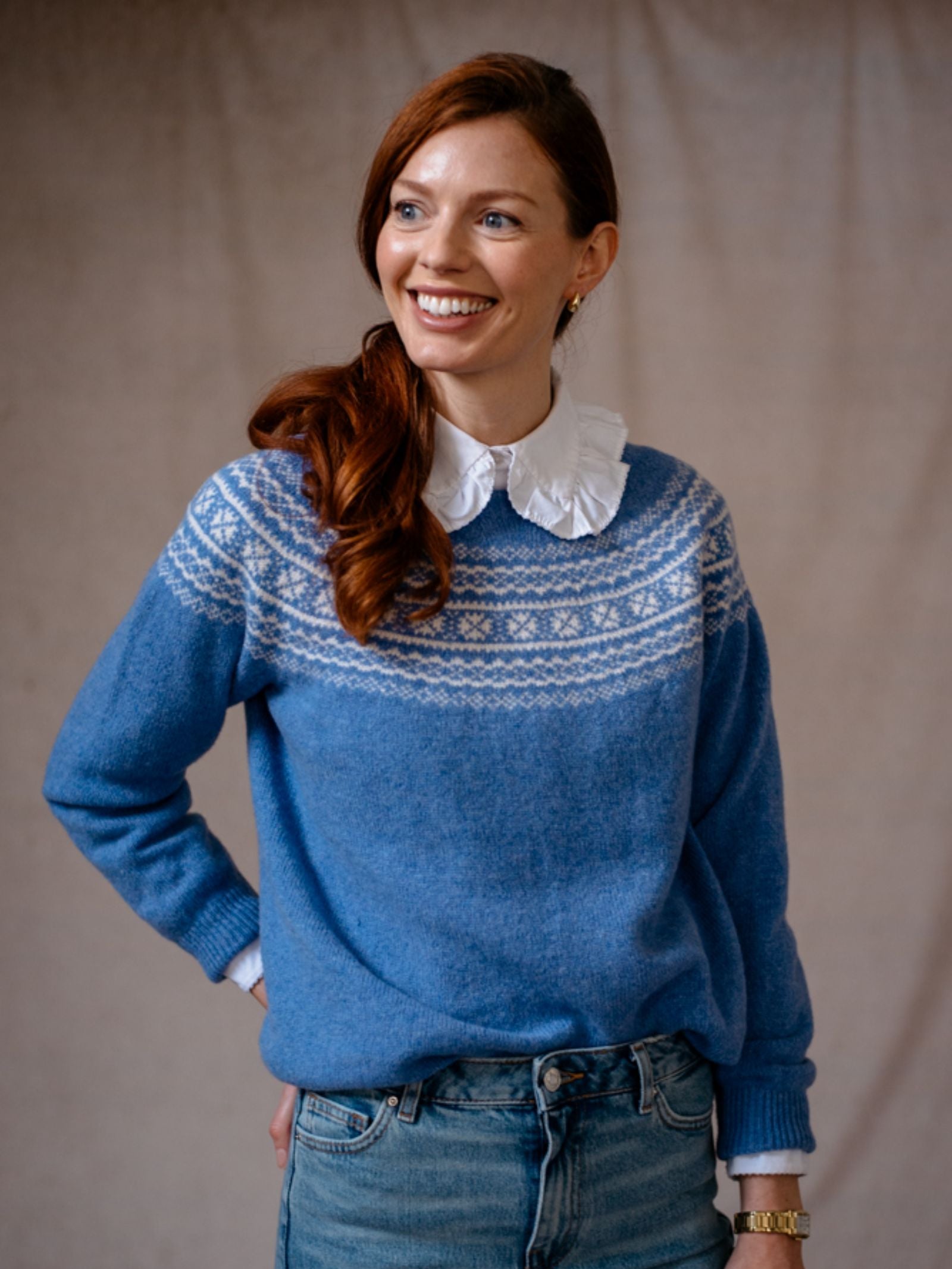 A woman with long red hair styled to one side smiles and looks left, wearing a Campbells of Beauly Two-Colour Fairisle Yoke Jumper over a white collared shirt and light blue jeans. The background is plain beige fabric.