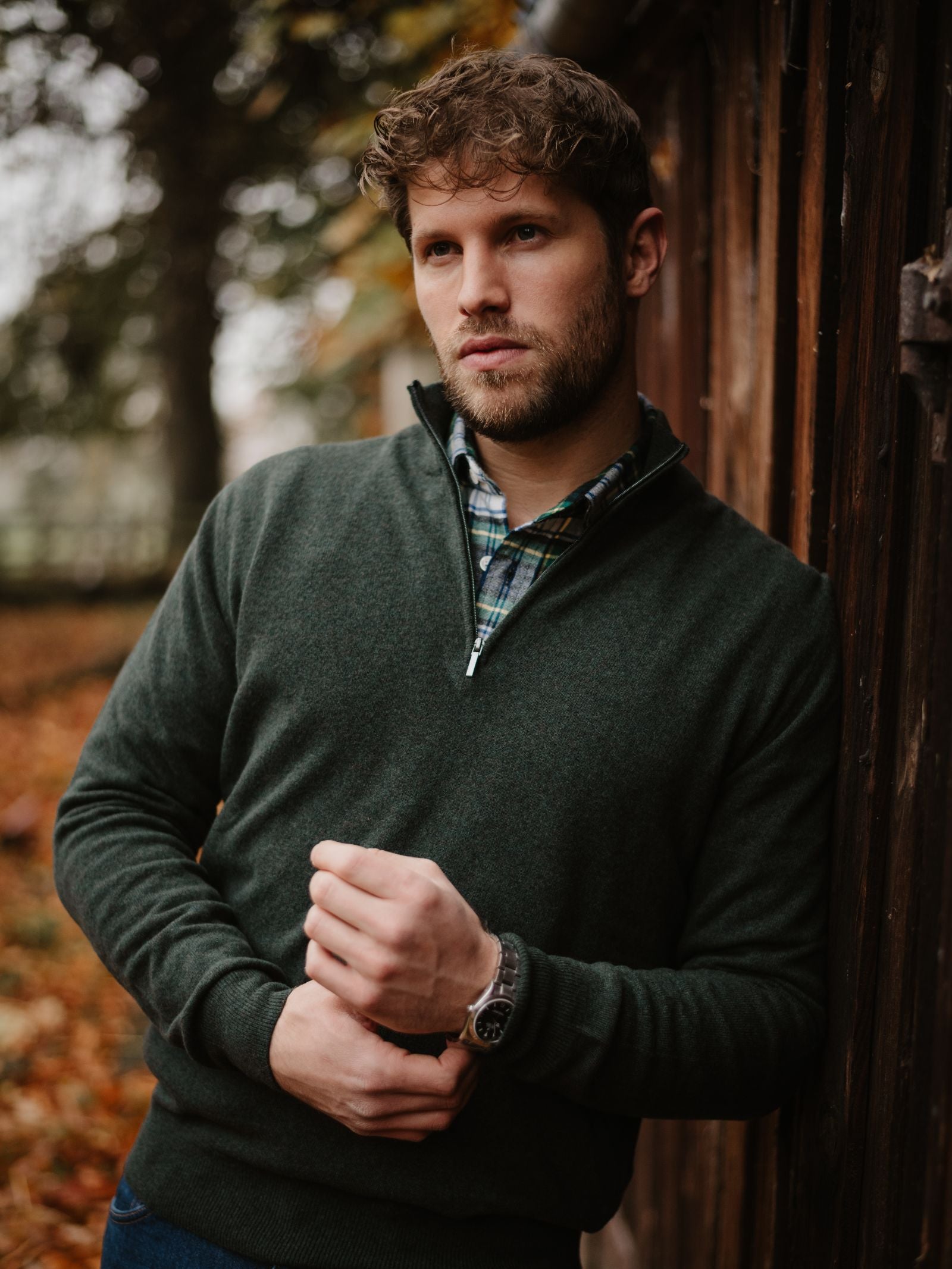 A man with curly hair and a beard stands outdoors by a wooden wall, wearing a Campbell's of Beauly Merino & Cashmere Quarter Zip Jumper over a plaid shirt. Blurred autumn leaves and trees are in the background.