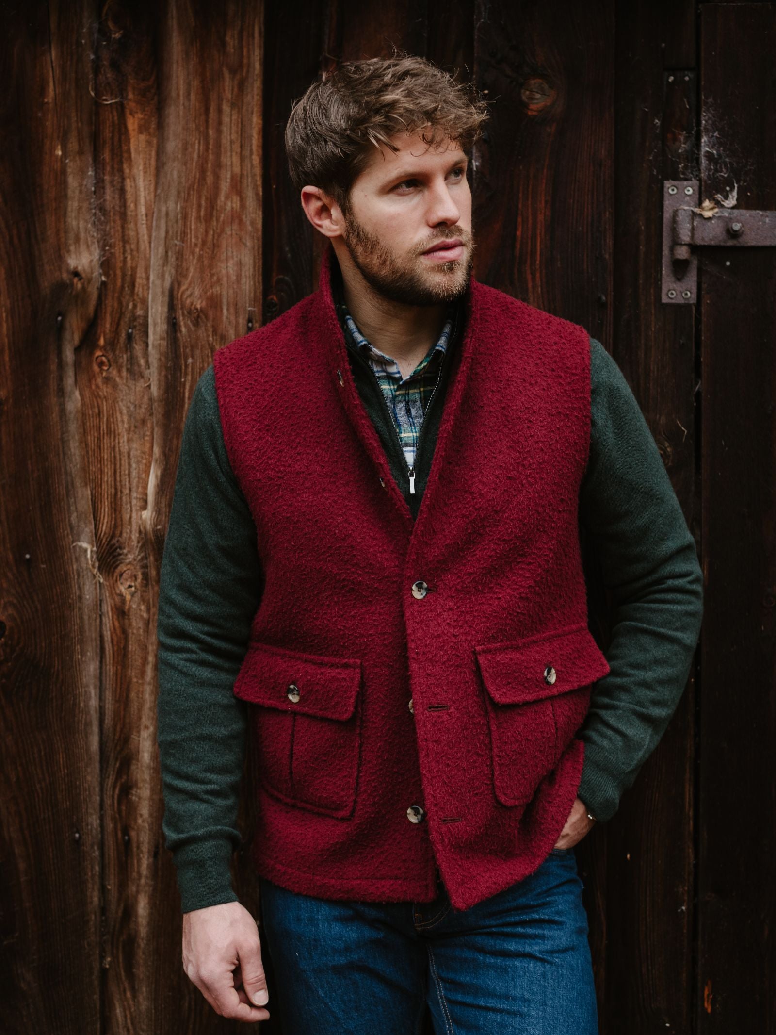 A man with short brown hair and a beard wears a red Campbell's of Beauly Casentino Fleece Gilet over a green shirt and blue jeans, standing before a rustic wooden wall and glancing to the side.