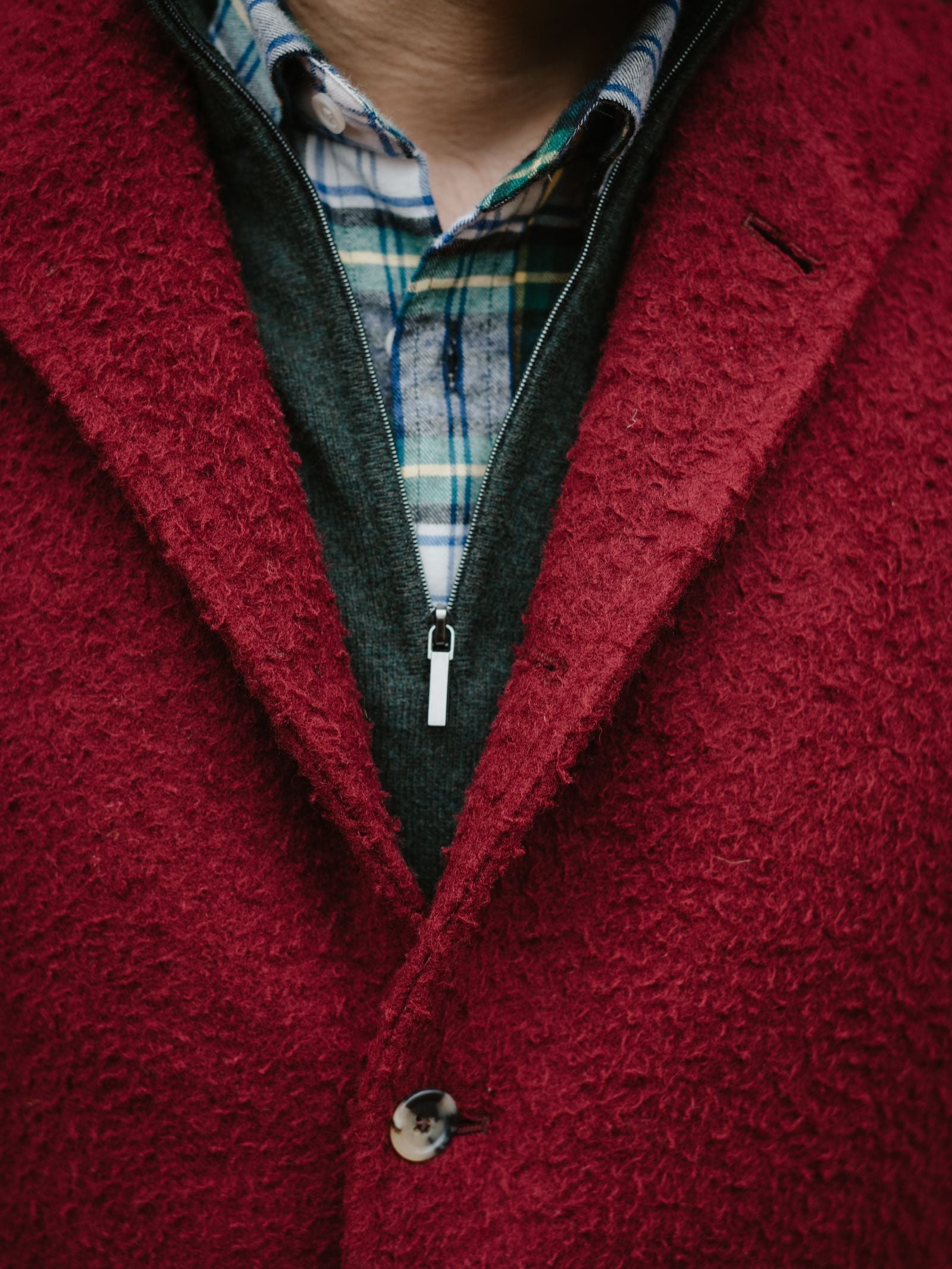 A close-up of a person wearing a red textured coat over the Campbell's of Beauly Casentino Fleece Gilet and a dark zip-up sweater with a blue plaid shirt, part of their neck and chin visible.