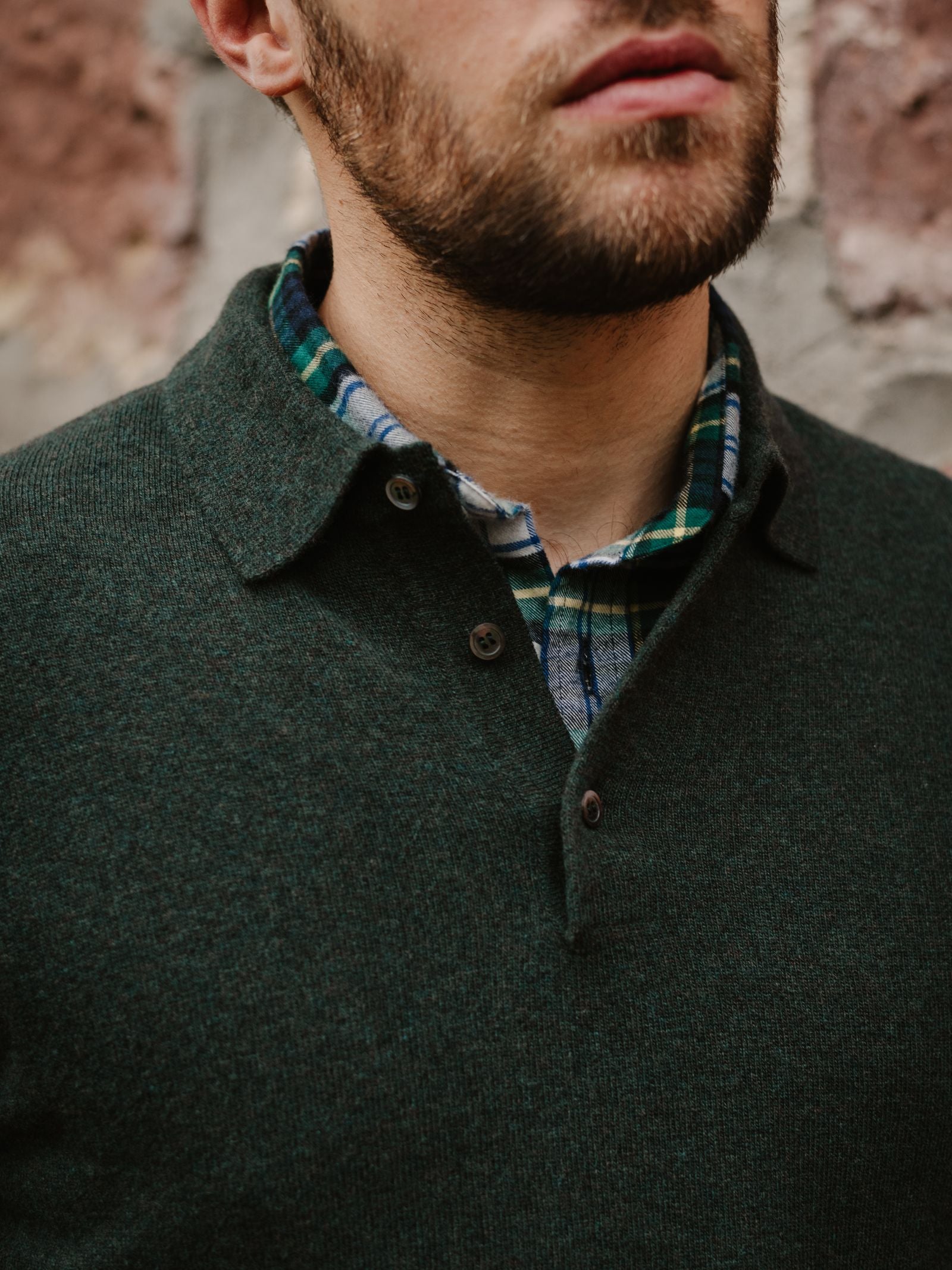 A bearded man wears the Campbell's of Beauly Merino & Cashmere Shirt Jumper in dark green, layered over a green and blue plaid shirt. He stands before a textured stone wall, shown from the nose down.