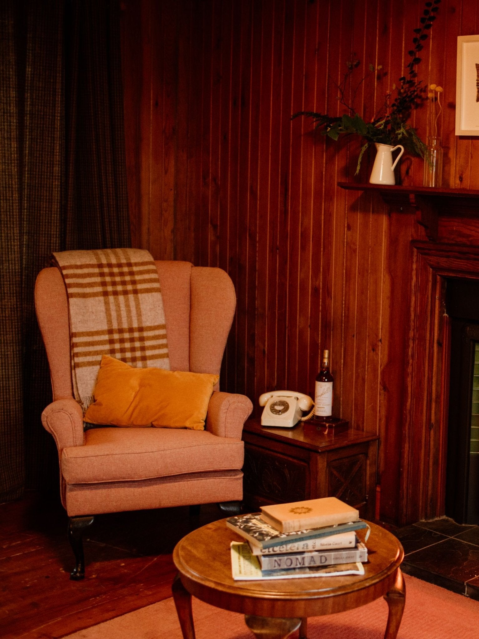 A cozy nook with a peach armchair draped in a Campbell's of Beauly Tarradale Lambswool Throw (check design) and orange cushion, next to a wooden table with vintage rotary phone, wine bottle, books, and wood-paneled walls for extra warmth.