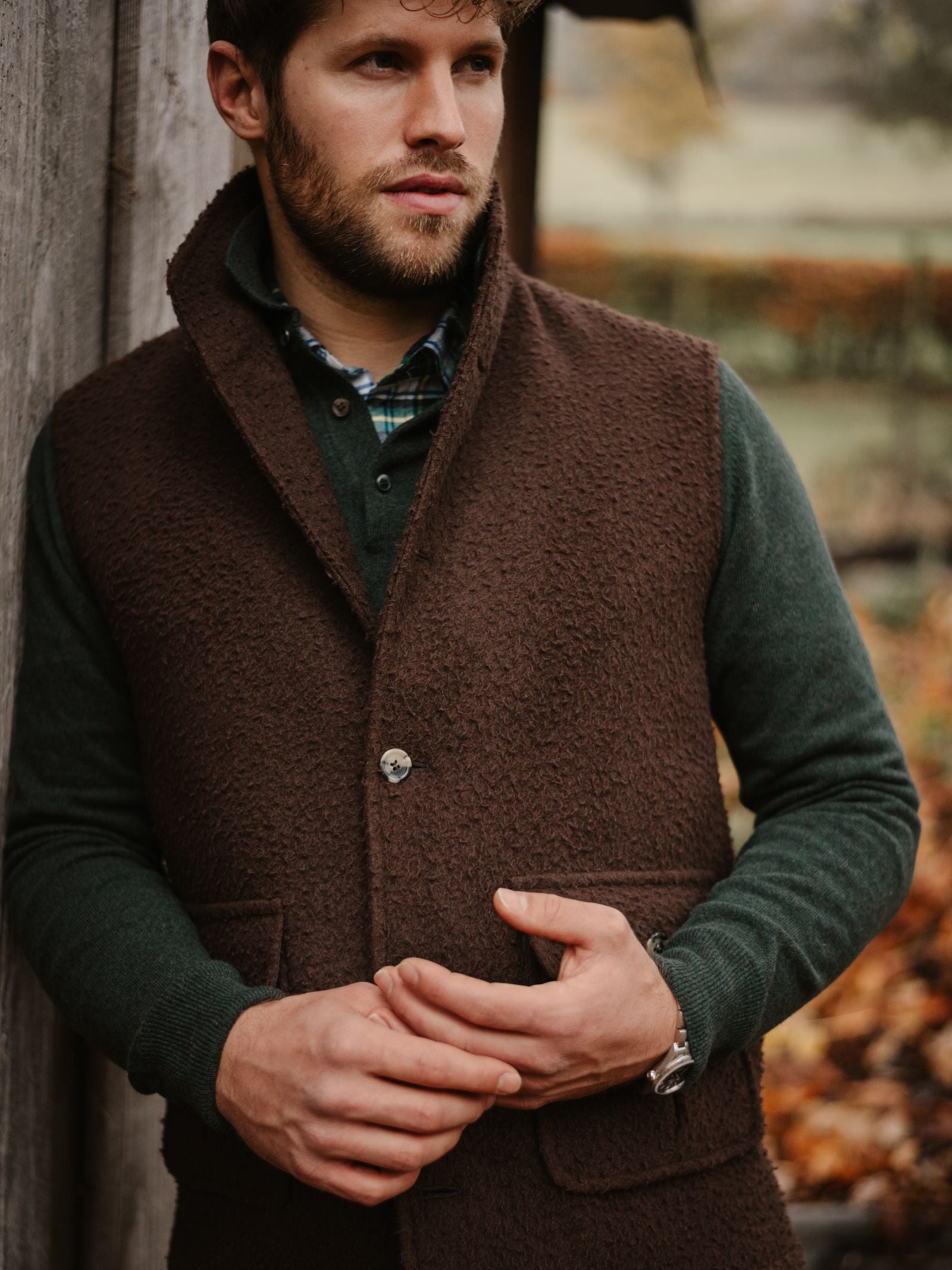 A bearded man stands outdoors in a dark green shirt and the Casentino Fleece Gilet by Campbell's of Beauly, leaning against a wooden wall with blurred autumn foliage behind him, hands clasped together in front.