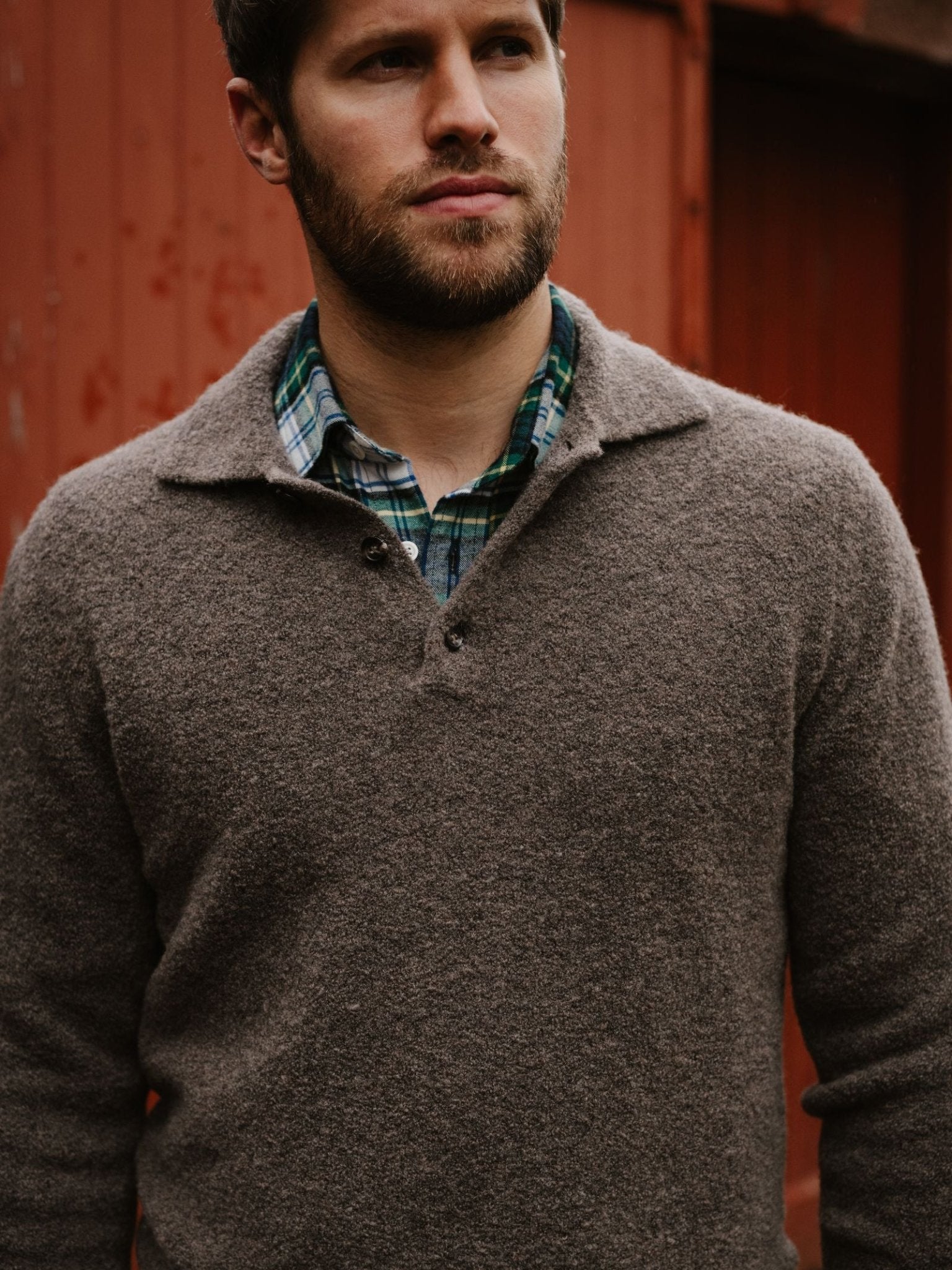 A bearded man wears the Campbell's of Beauly Bouclé Polo Sweater over a green plaid shirt, standing before a weathered red wooden wall.