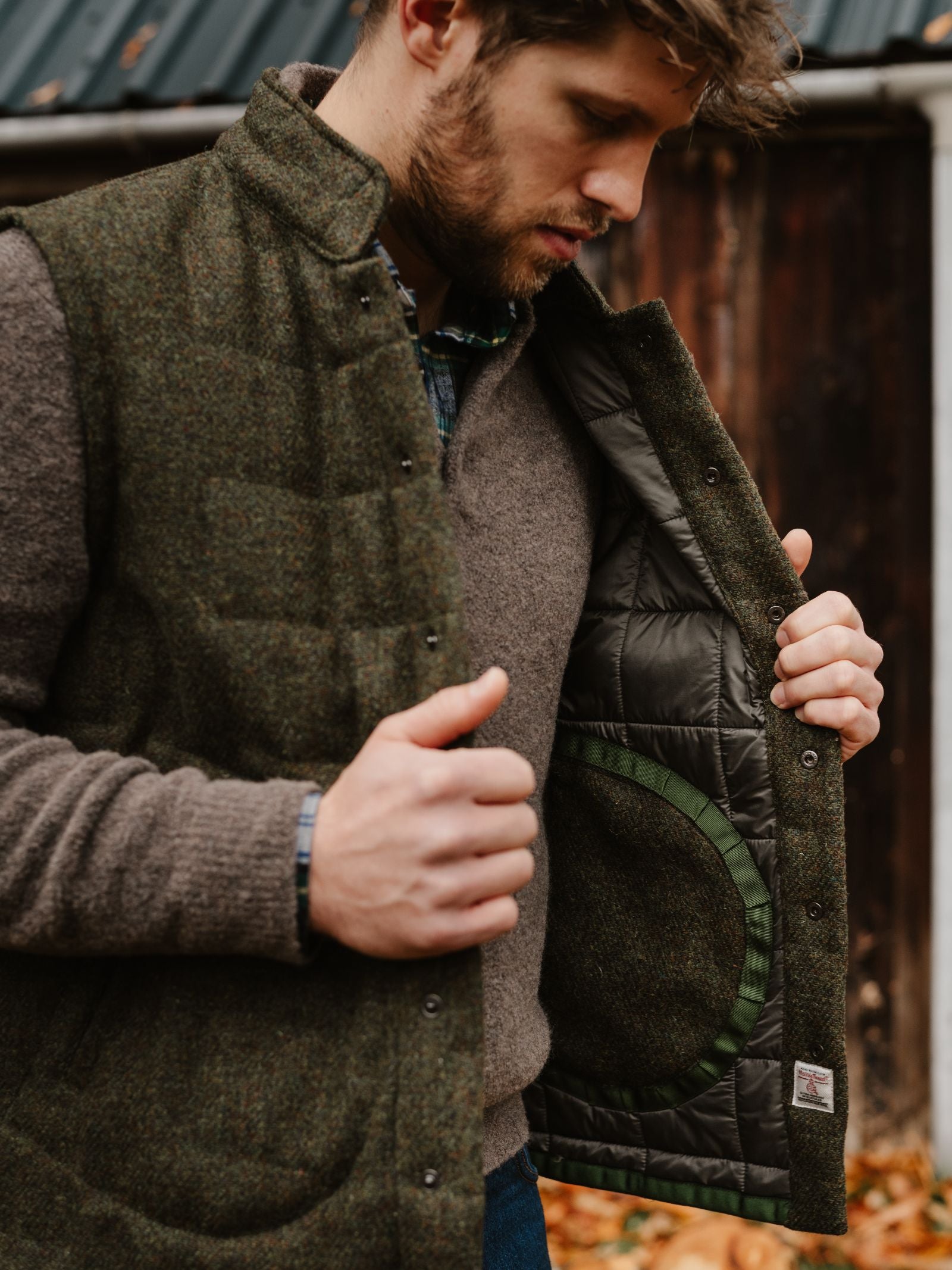 A man outdoors holds open his Campbell's of Beauly Harris Tweed Quilted Gilet, showing its quilted lining. He wears a brown wool sweater; autumn leaves and a wooden building are in the background.