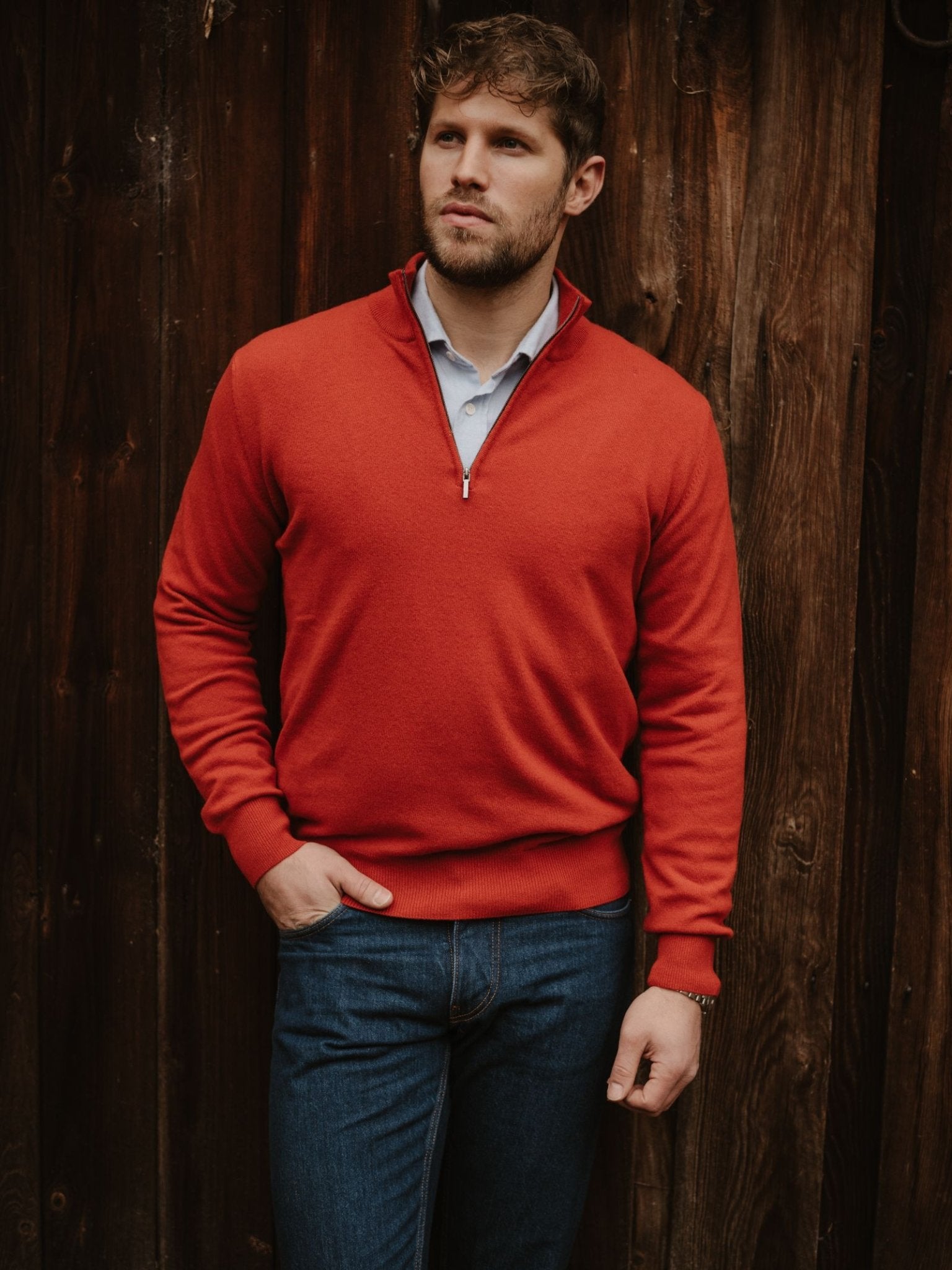 A man with short brown hair and a beard stands by a wooden wall, wearing Campbell's of Beauly Merino & Cashmere Quarter Zip Jumper over a light blue shirt and blue jeans, one hand in his pocket, looking to the side.