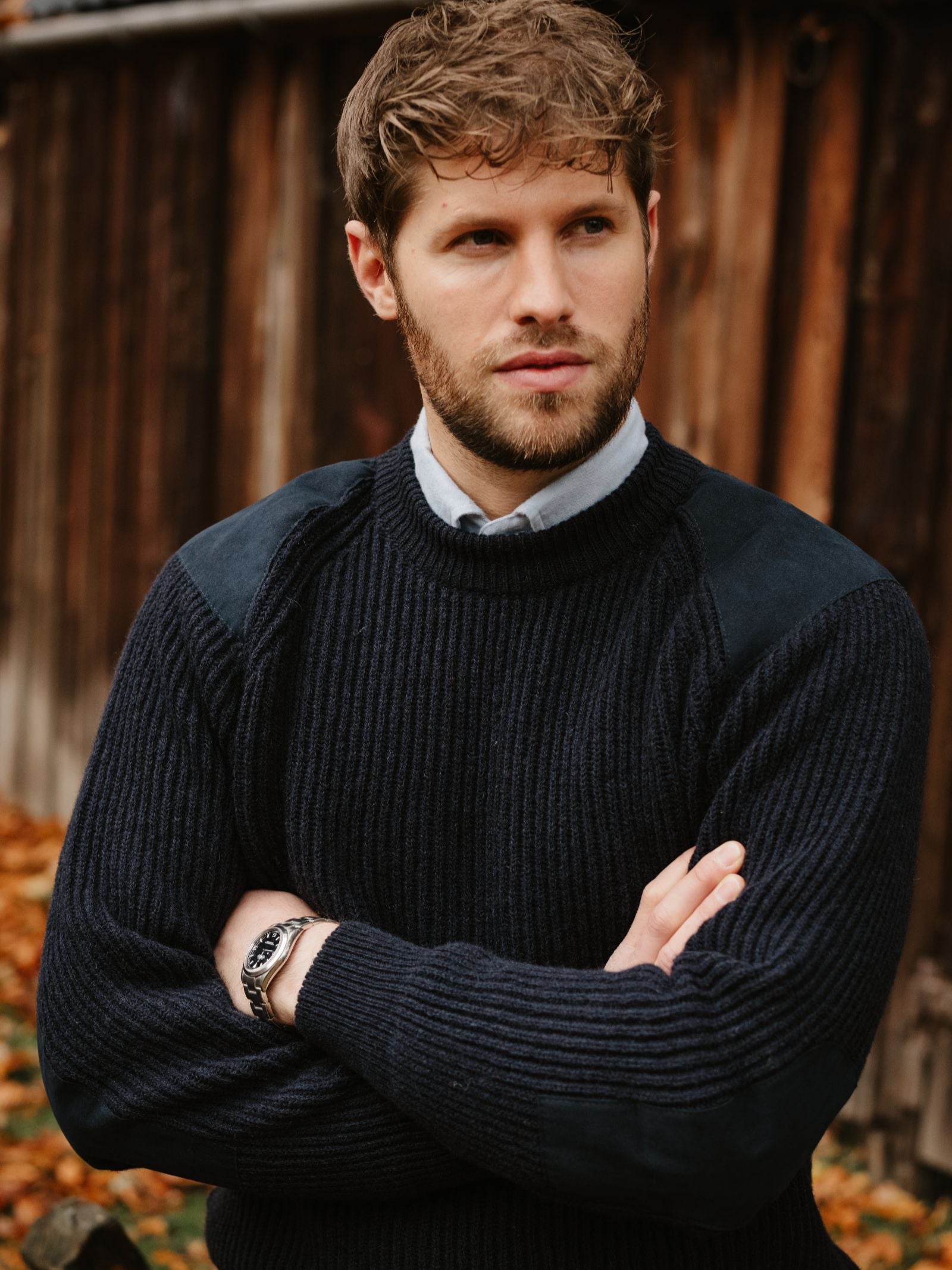 A man with light brown hair and a beard stands outdoors with his arms crossed, wearing the Scotia Suede Jumper from Campbell's of Beauly over a collared shirt. Wooden planks and autumn leaves are in the background.