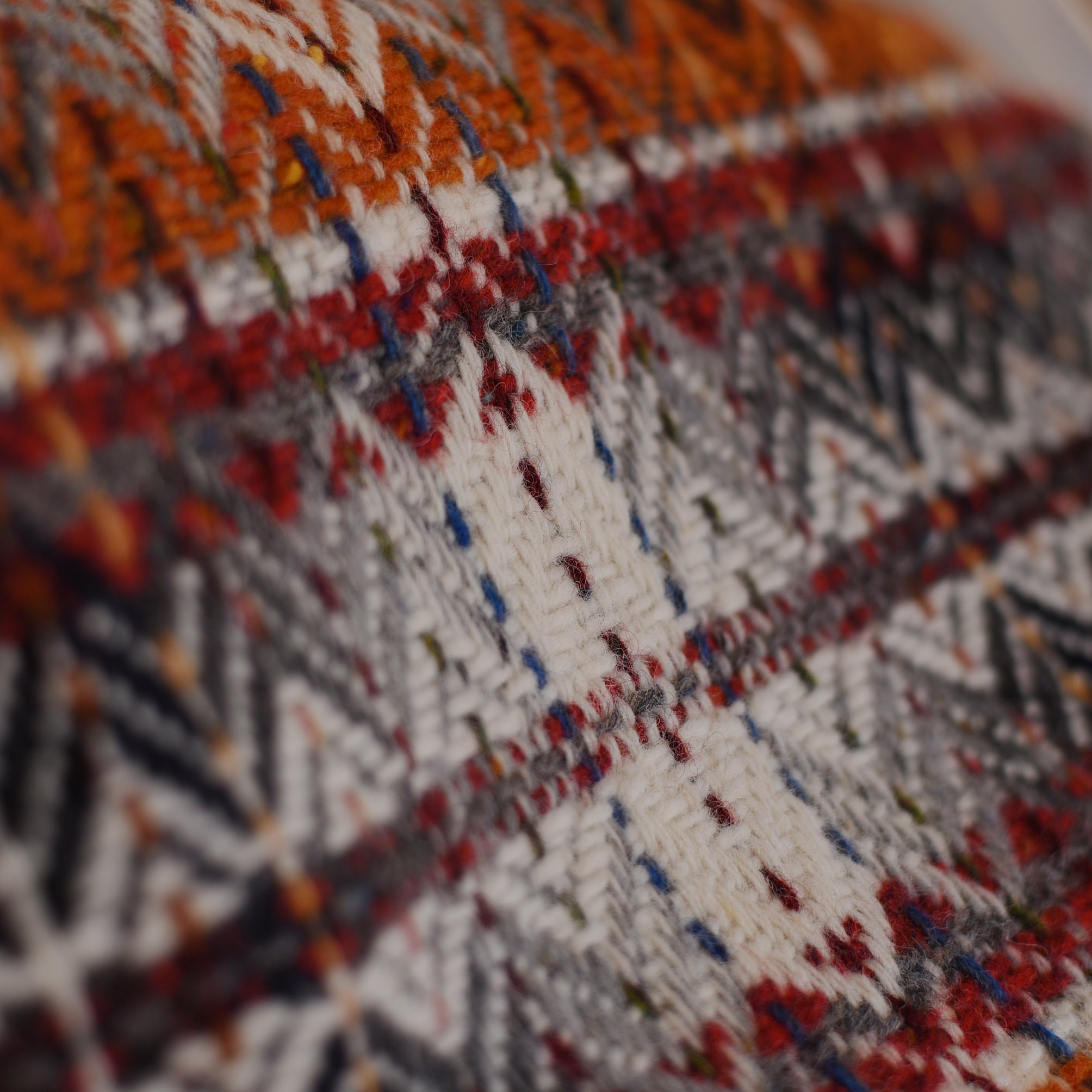 Close-up of the Wyvis Lambswool Throw by Campbells of Beauly, showcasing intricate geometric patterns and textured stitches in orange, red, gray, white, and blue flecked yarns, reminiscent of traditional diamond weave.