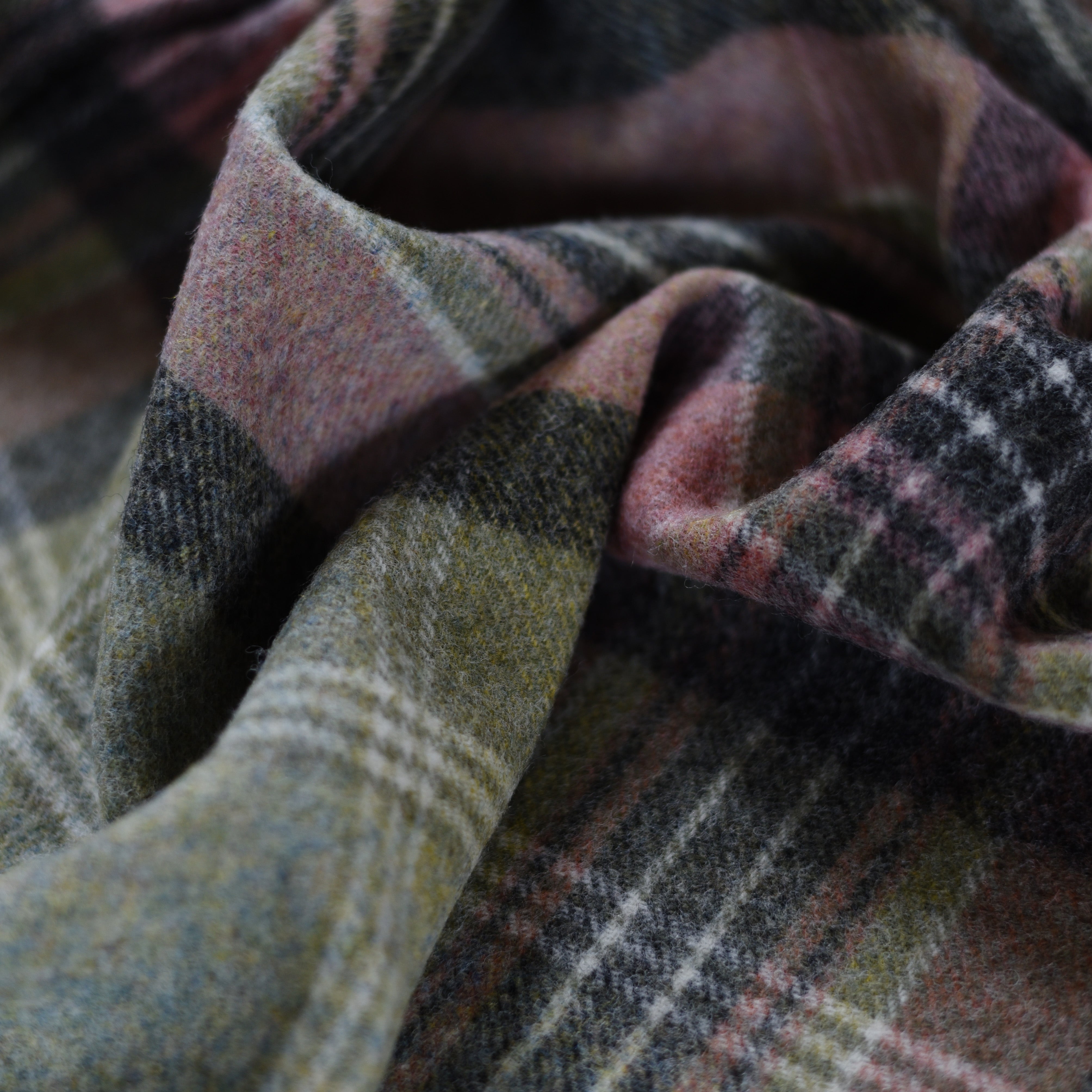 A close-up of the Inchmore Lambswool Throw from Campbells of Beauly shows soft, folded plaid fabric in muted green, grey, pink, and cream. The Yorkshire lambswool appears textured and cozy with gentle folds and inviting shadows.
