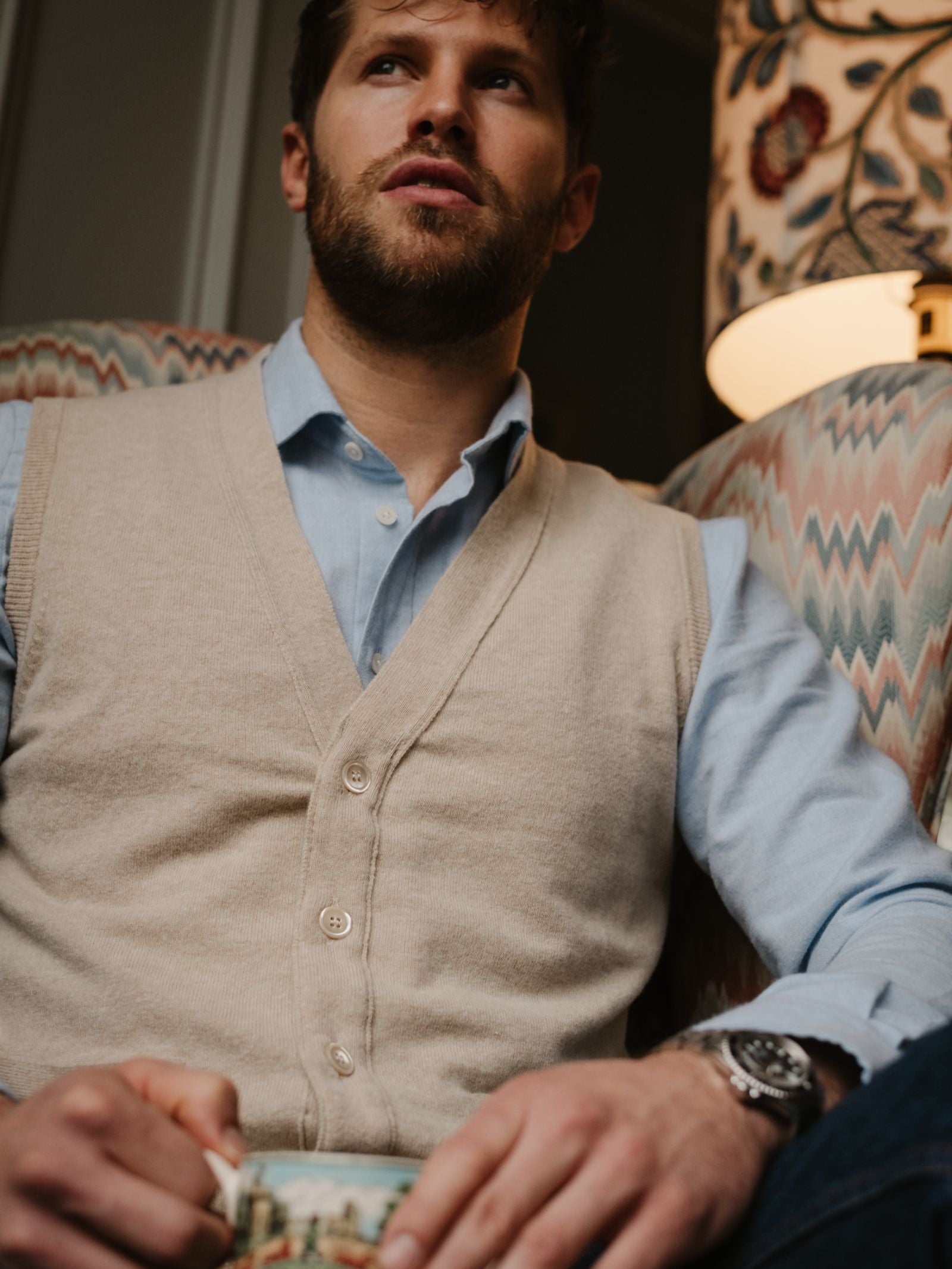 A man with a trimmed beard sits in an armchair, wearing a light blue shirt and the Campbell's of Beauly Geelong Lambswool Button Waistcoat, holding a book. A patterned lampshade and armchair complete this cozy country wardrobe scene.