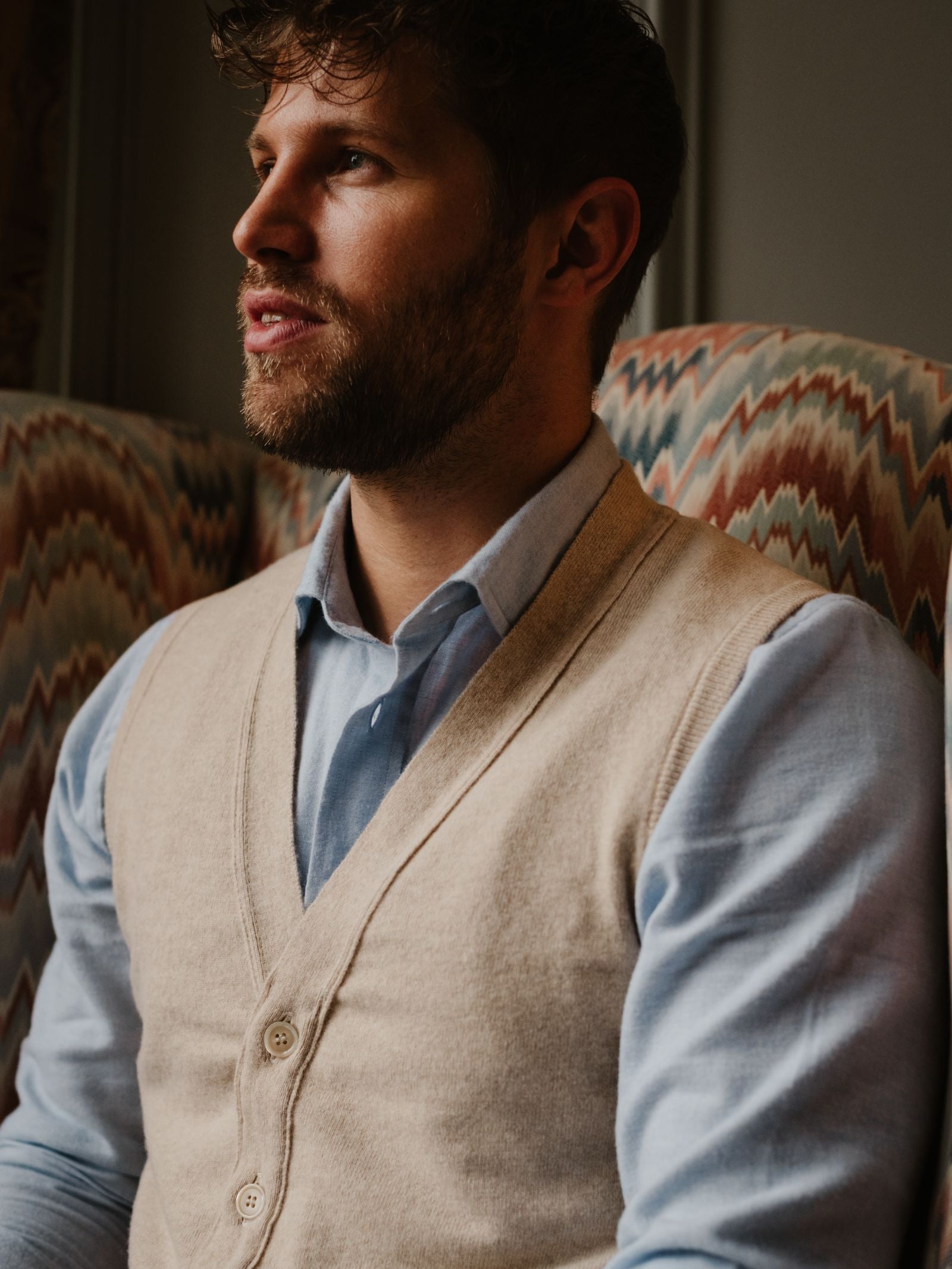 A man with wavy brown hair and a beard sits on a patterned chair, wearing a light blue shirt and the Campbell's of Beauly Geelong Lambswool Button Waistcoat—an elegant choice for any country wardrobe.