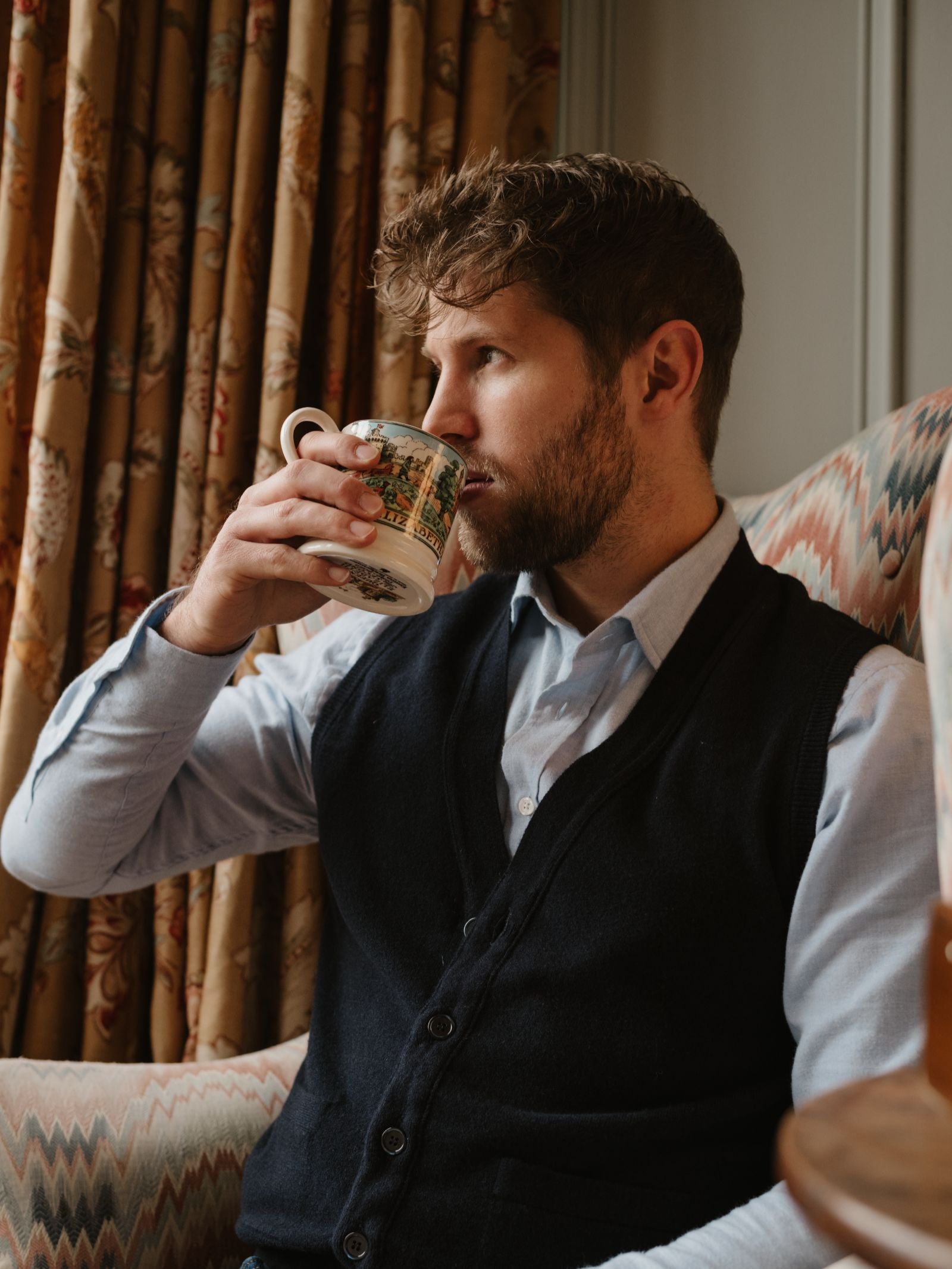 A man with short brown hair and a beard sits in an armchair, wearing a light blue shirt and the Campbell's of Beauly Geelong Lambswool Button Waistcoat, sipping from a patterned mug. Patterned curtains hang behind him.