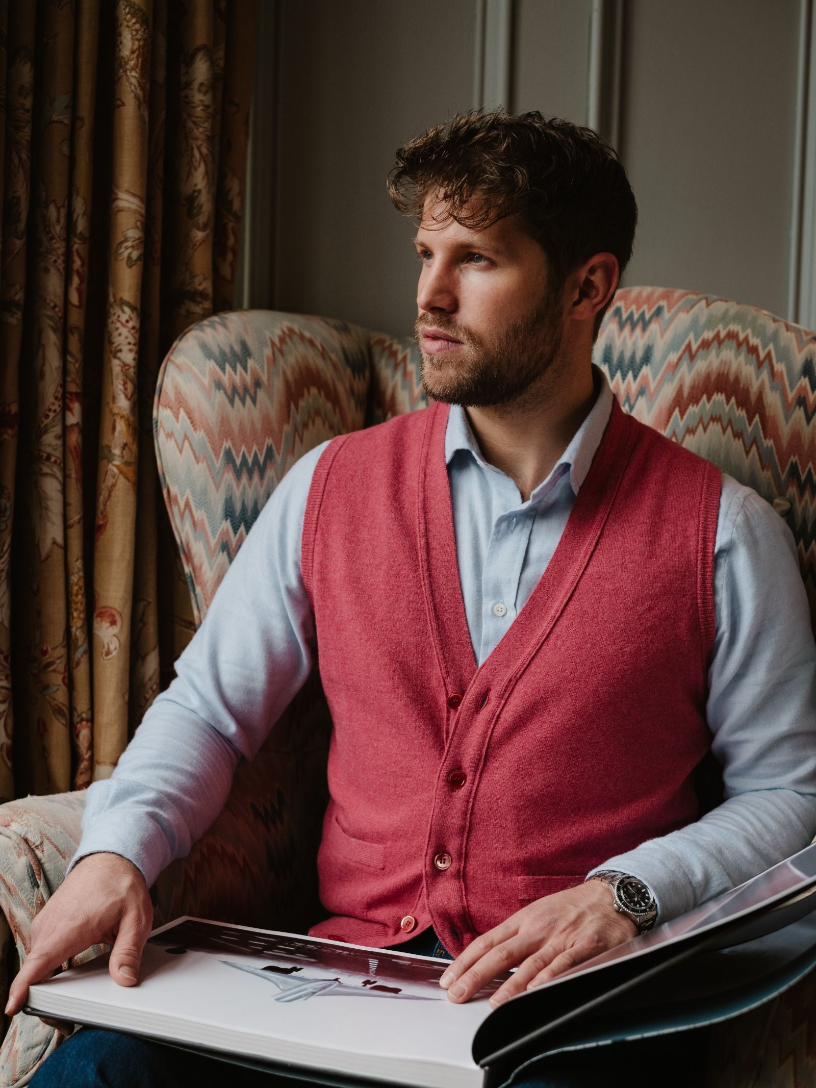 A man with curly hair, dressed in a light blue shirt and a Campbell's of Beauly Geelong Lambswool Button Waistcoat, sits in an armchair with an open book on his lap, looking thoughtfully out of frame. Patterned curtains and an upholstered chair are behind him.