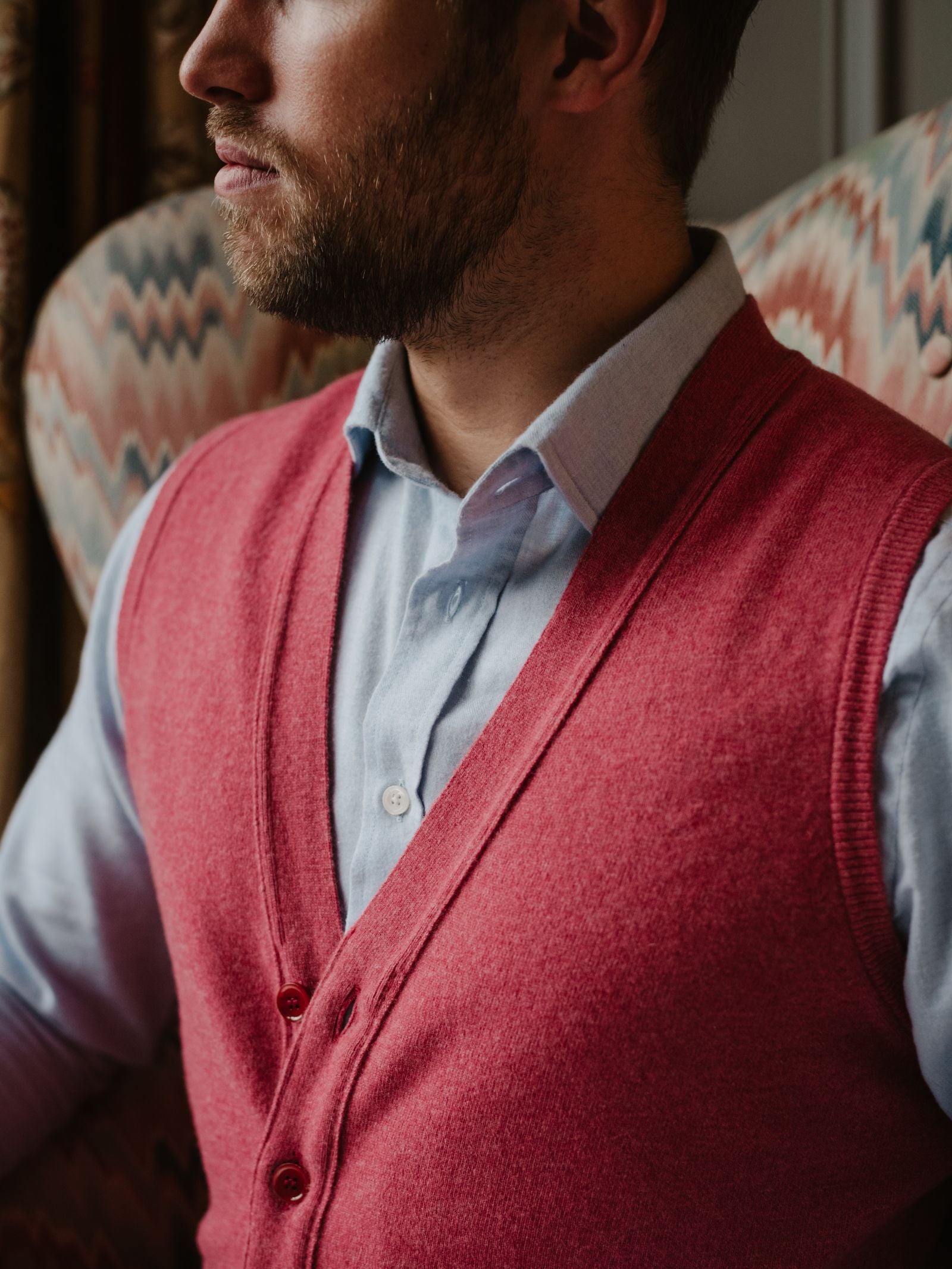 A bearded man in a light blue collared shirt and a red Campbell's of Beauly Geelong Lambswool Button Waistcoat sits before a patterned backdrop, his profile partly visible, capturing classic country wardrobe style.
