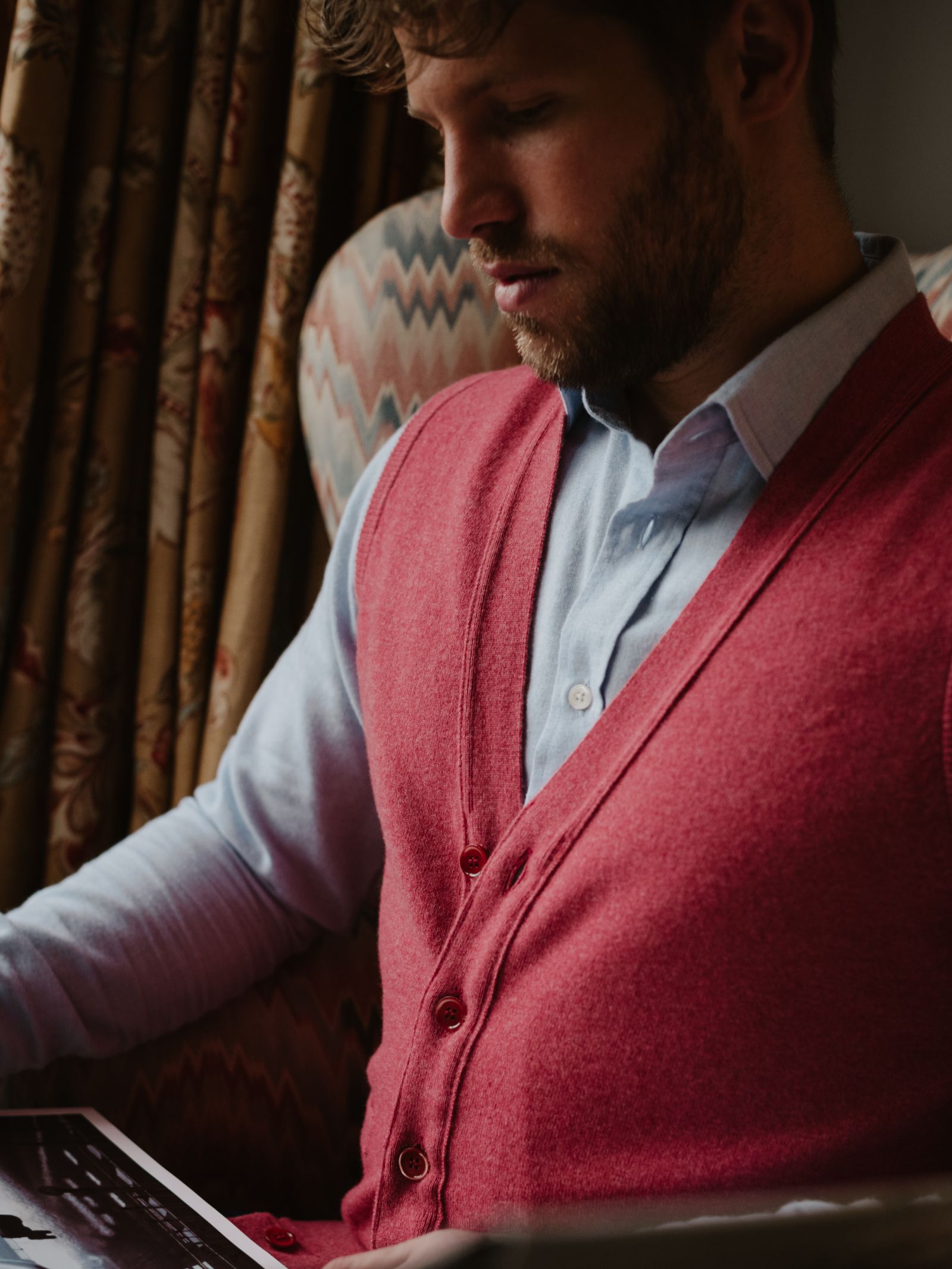 A bearded man wearing a Campbell's of Beauly Geelong Lambswool Button Waistcoat sits in a patterned armchair by a window with draped curtains, looking down at something in his hands.