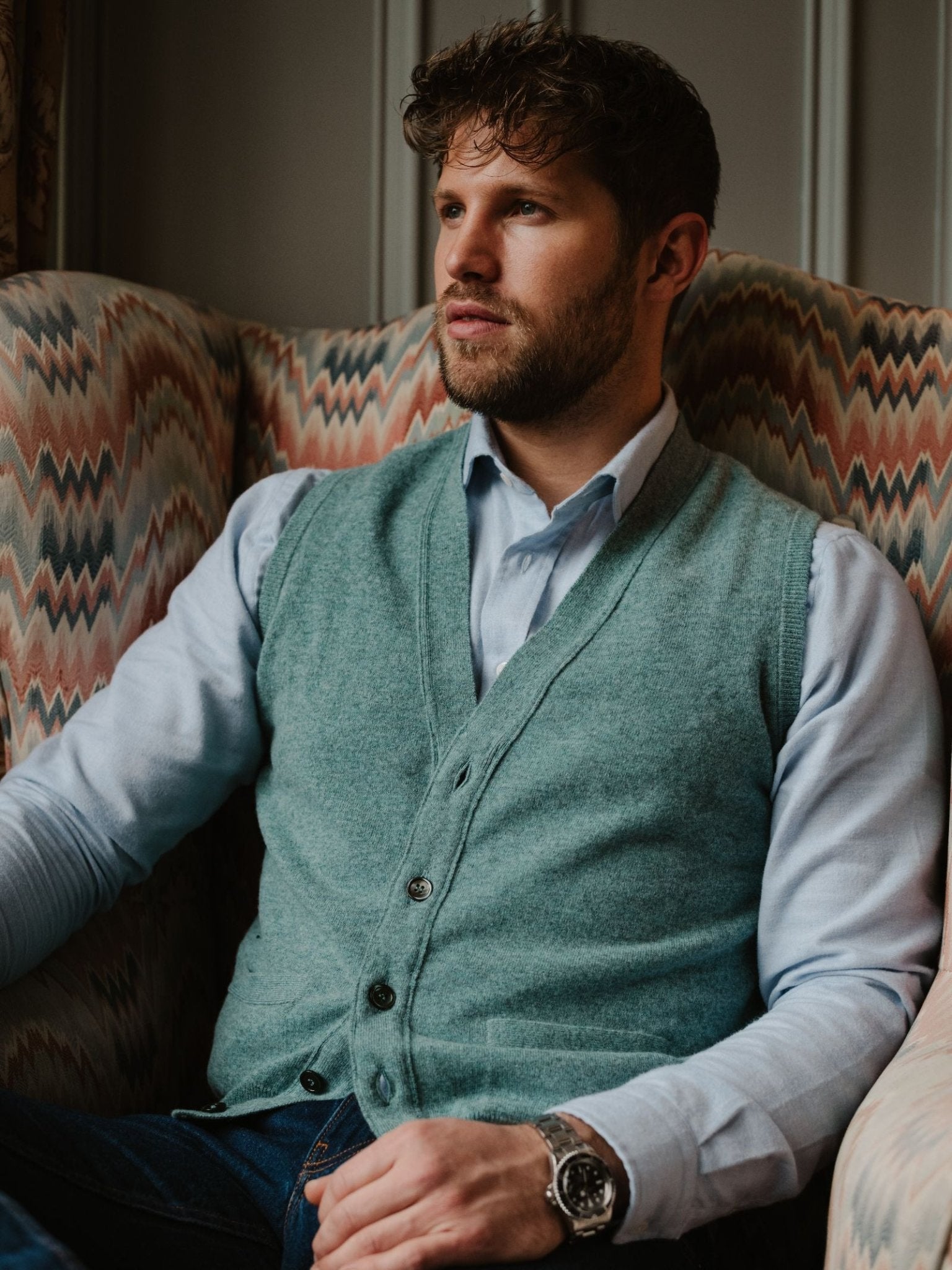A man with short, wavy brown hair and a beard sits in a patterned armchair, wearing a pale blue shirt and a Campbell's of Beauly Geelong Lambswool Button Waistcoat in light green, paired with a wristwatch, gazing thoughtfully to his left.