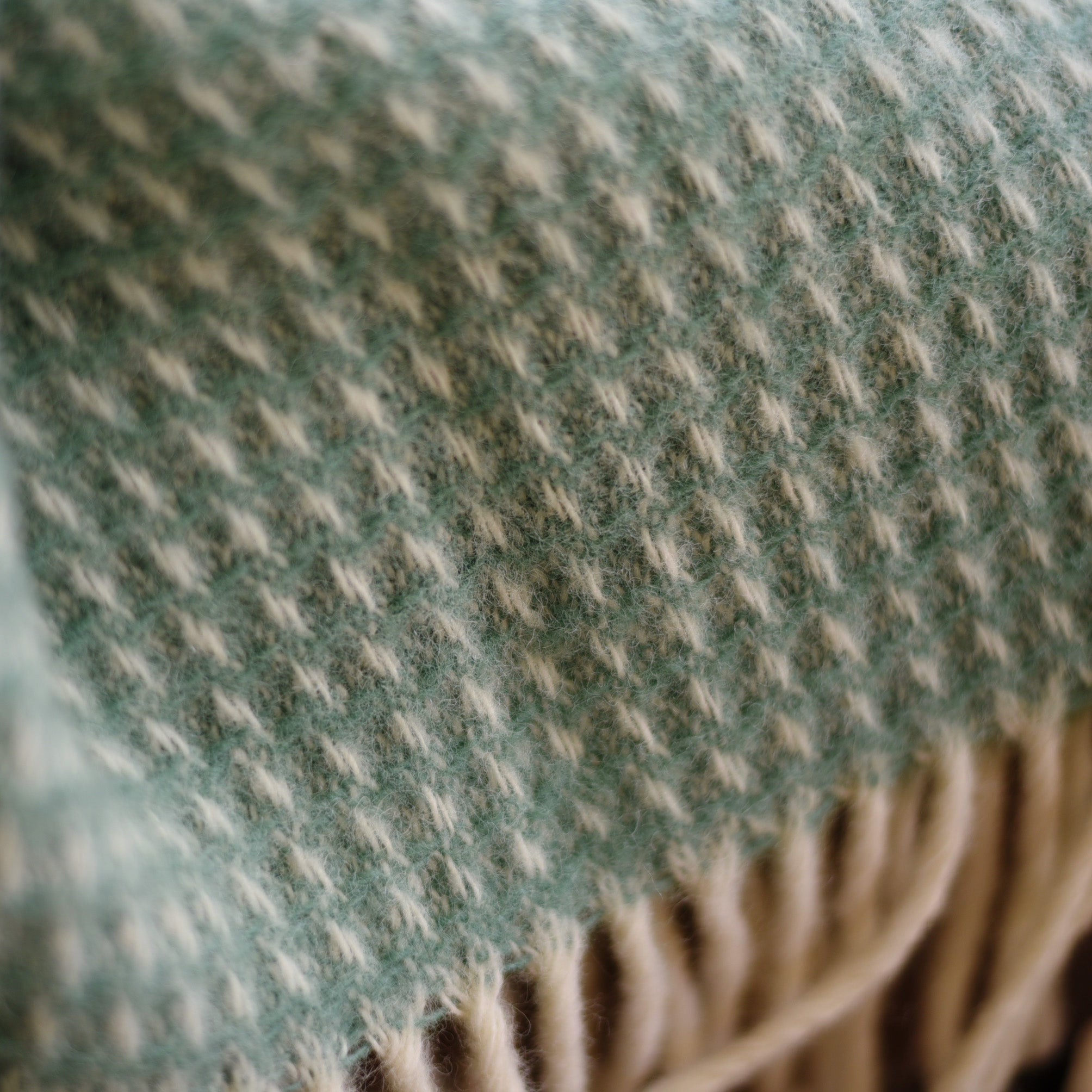 Close-up of a soft woven fabric featuring a light green and cream houndstooth pattern with beige fringe at the edge, from the Honeycomb Lambswool Throw by Campbells of Beauly.