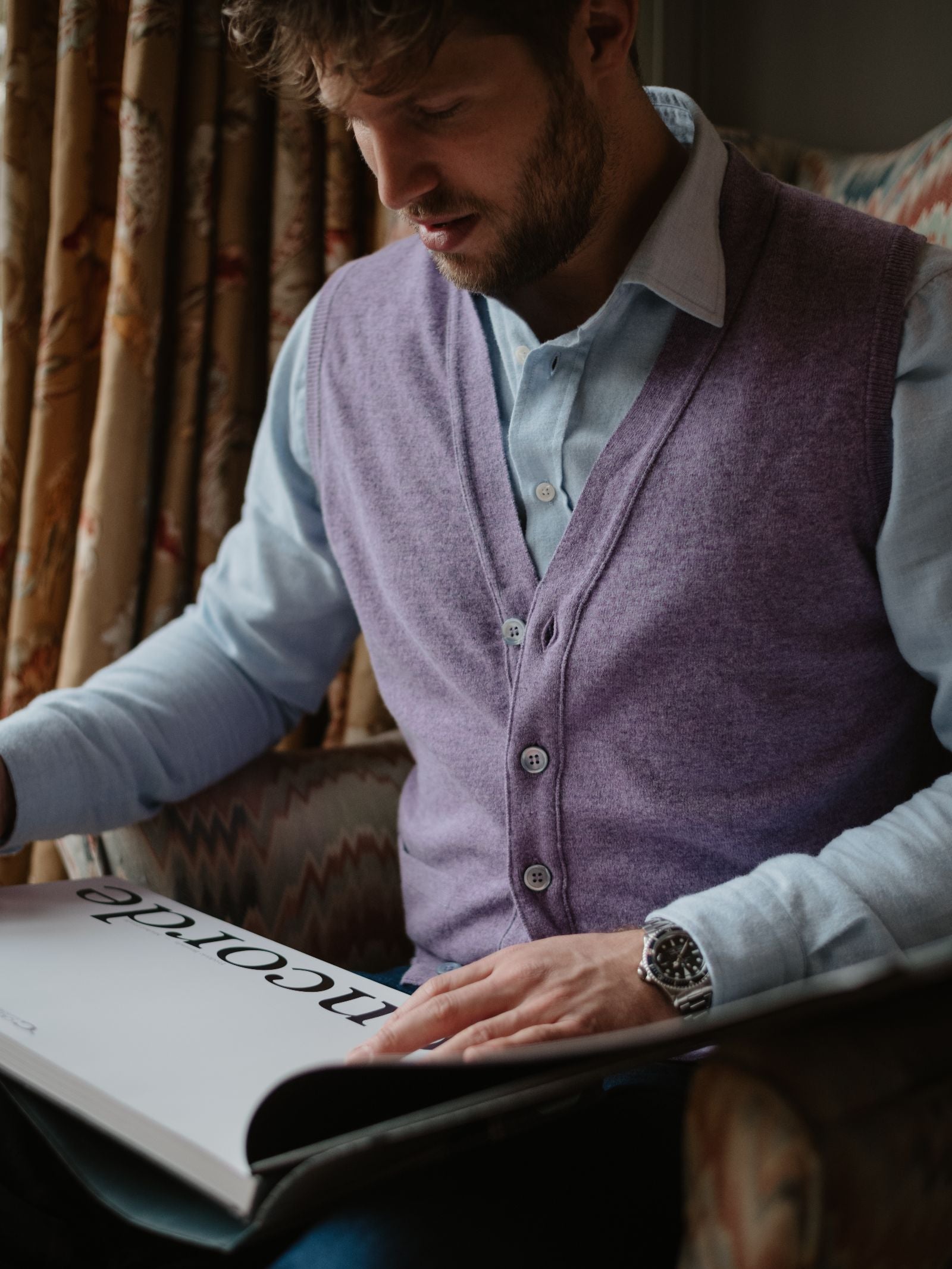 A man in a light blue shirt and a Campbell's of Beauly Geelong Lambswool Button Waistcoat sits in an armchair, engrossed in a large book titled PICOTADO, surrounded by patterned upholstery and country-style curtains.