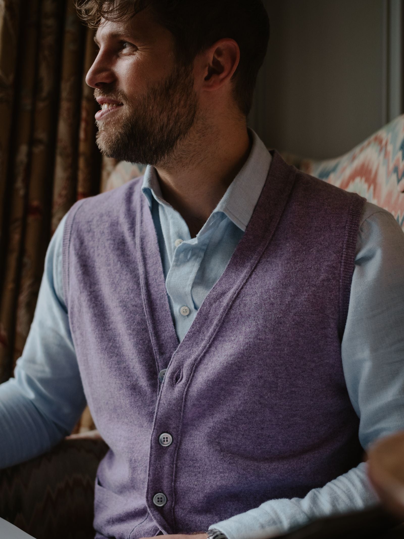 A bearded man in a light blue button-up and Campbell's of Beauly Geelong Lambswool Button Waistcoat sits in a patterned armchair, gazing sideways with a slight smile. Soft lighting and curtains complete the refined country wardrobe setting.
