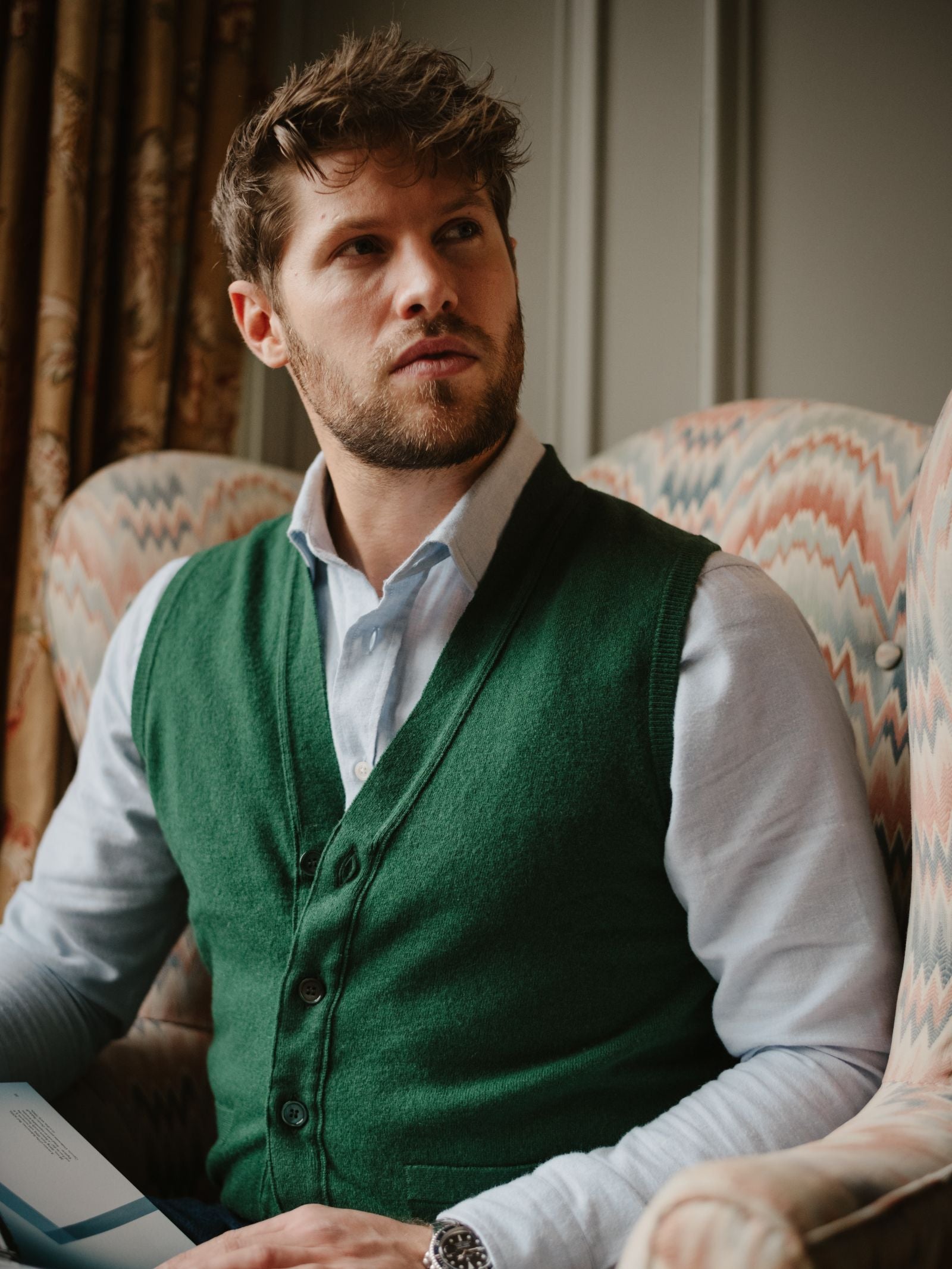 A man with light brown hair and a beard sits in an armchair, wearing a light blue shirt and the Geelong Lambswool Button Waistcoat by Campbell's of Beauly. He gazes upward, holding a booklet, with patterned curtains adding to the country wardrobe vibe.