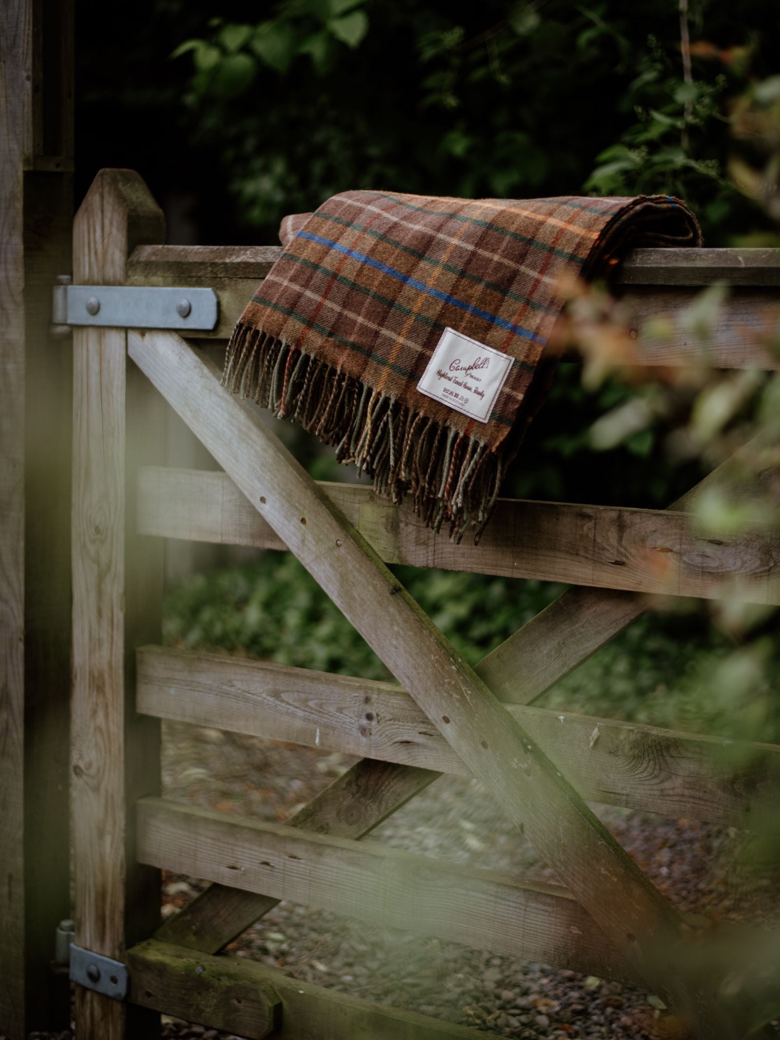 A Lavender Throw by Campbells of Beauly with fringed edges hangs over a rustic wooden gate outdoors, featuring metal hardware and a gravel path, with green foliage and trees softly blurred in the background.