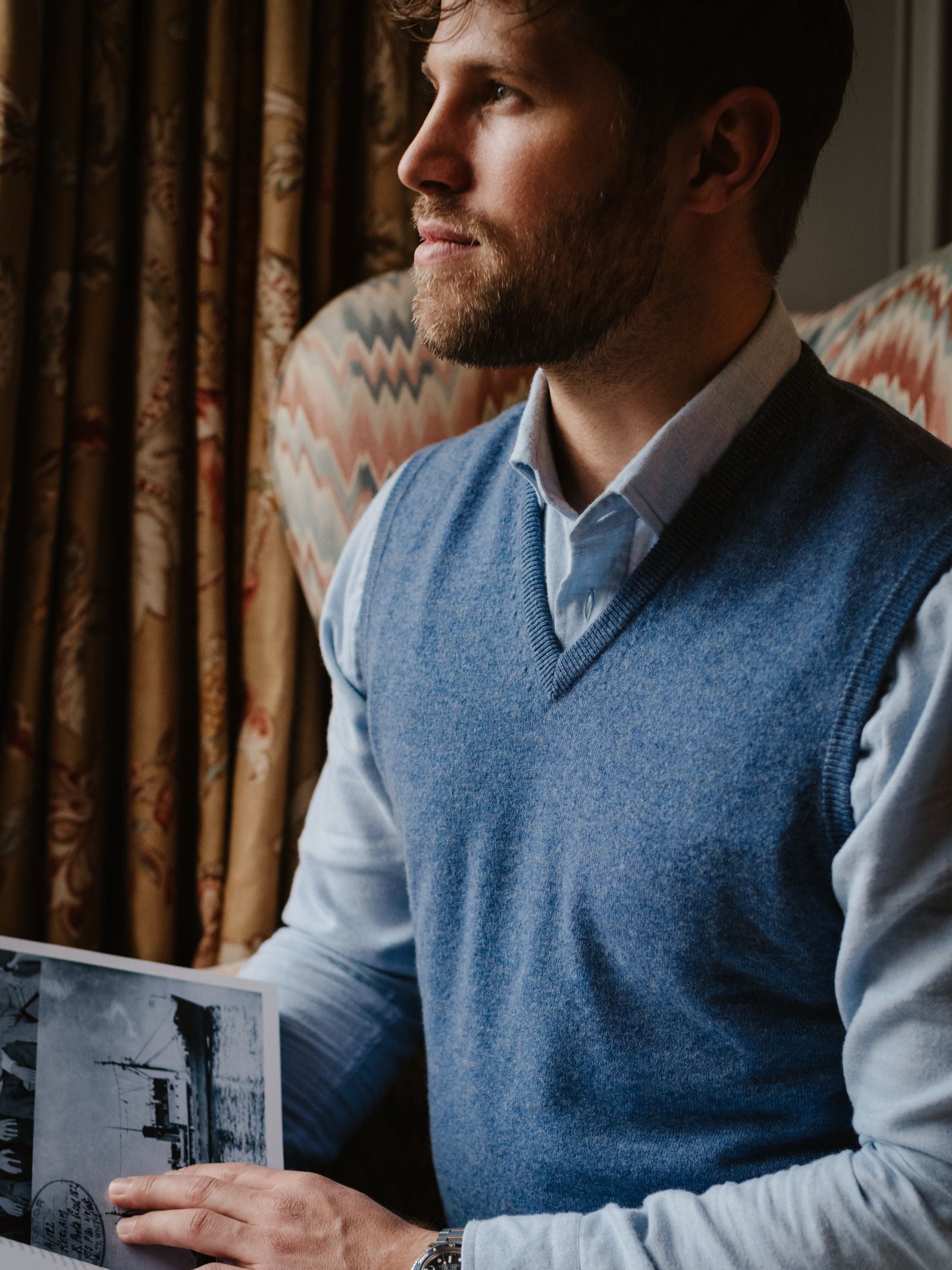 A bearded man in a classic-fit Campbell's of Beauly Cashmere Slipover over a light blue shirt sits by a window in a patterned chair, holding a black-and-white photo and gazing thoughtfully into the distance.