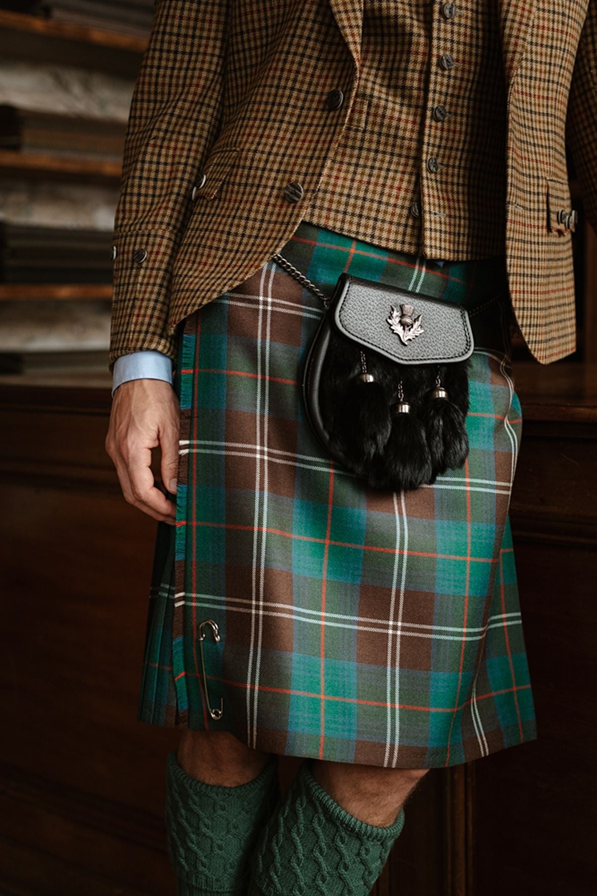 A person wearing a brown patterned jacket, a green and blue tartan kilt with a safety pin, and a black sporran.