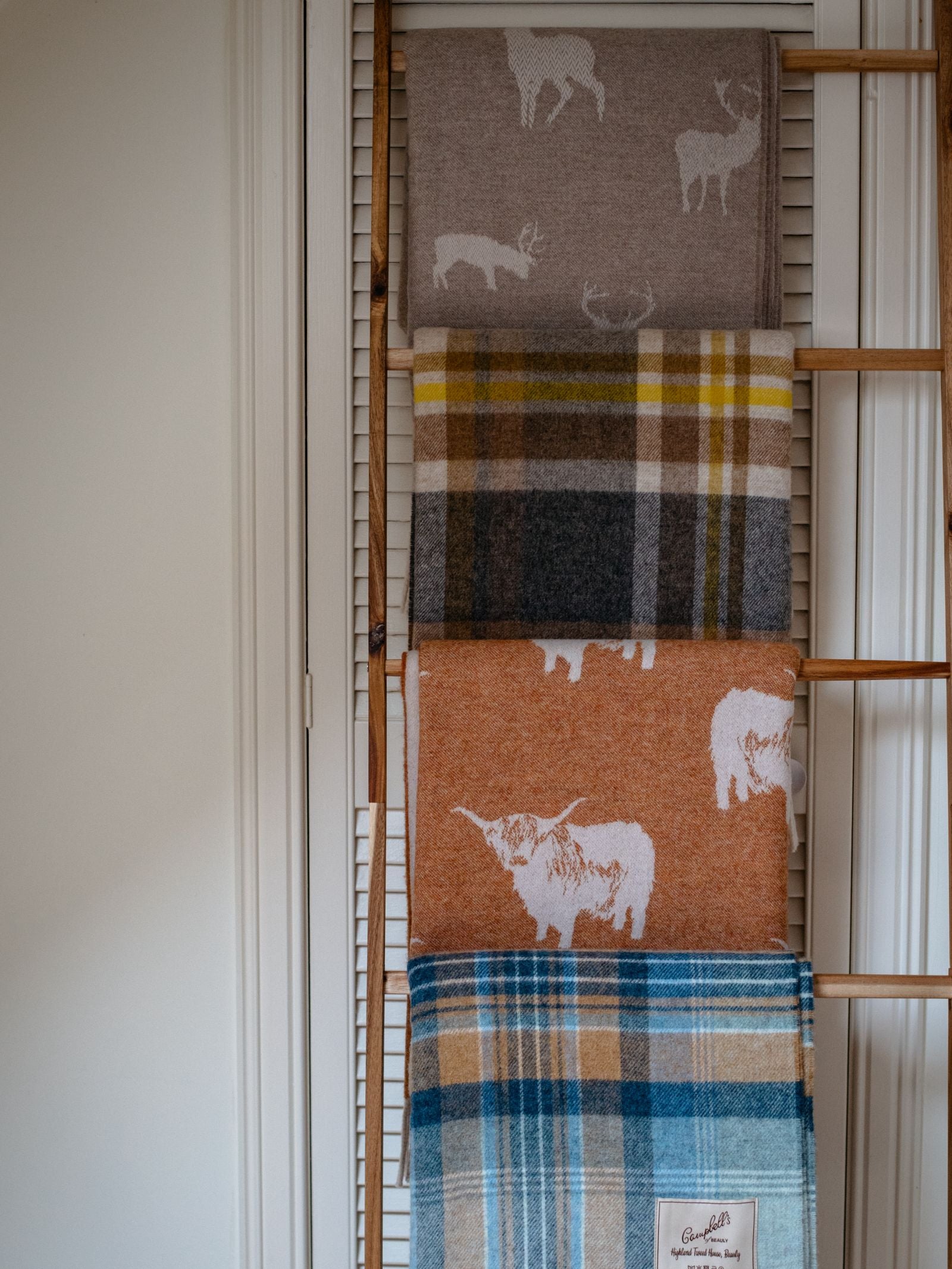 A wooden ladder leans against a white paneled wall, displaying four Campbells of Beauly Stag Lambswool Blankets—two with stag and cow motifs in beige/rust, and two plaid designs in blue, yellow, and gray—all beautifully woven in Scotland.