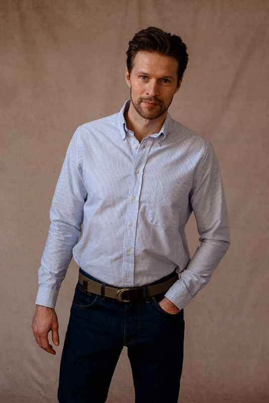 A man with short brown hair and light stubble stands against a plain beige backdrop, wearing a Campbells of Beauly Oxford Shirt tucked into dark blue jeans with a brown belt. One hand is in his pocket as he looks calmly at the camera.