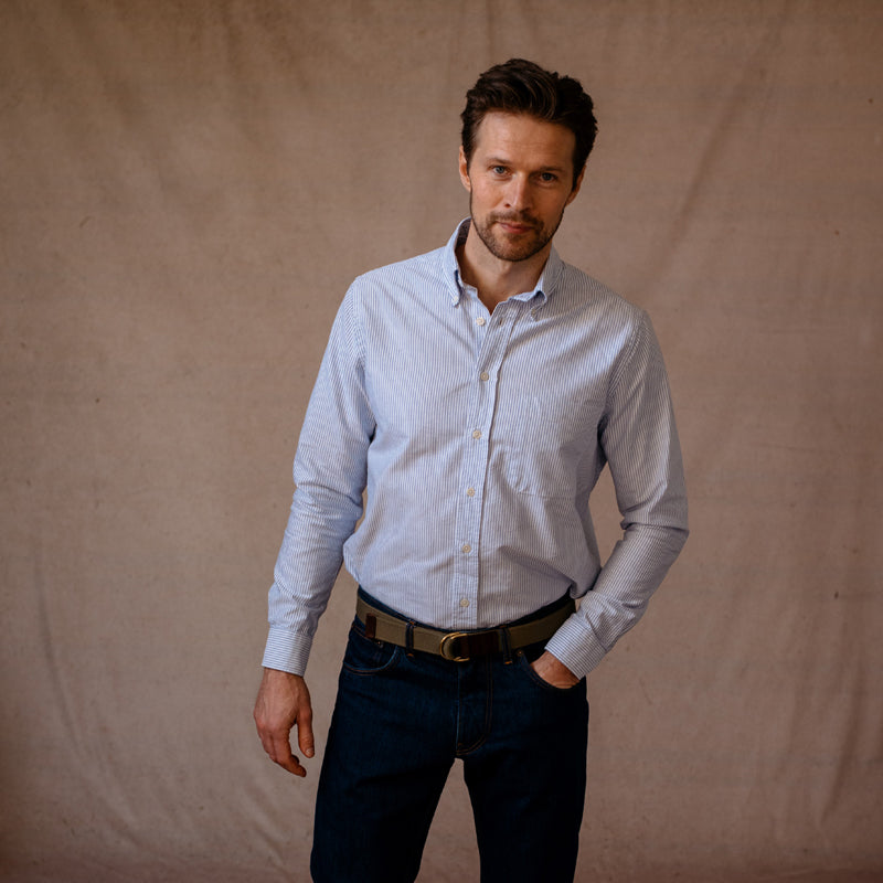 A man with short brown hair and a trimmed beard wears the Campbells of Beauly Oxford Shirt in light blue stripes, tucked into dark jeans with a brown belt. He stands against a beige textured background with one hand in his pocket and a neutral expression.
