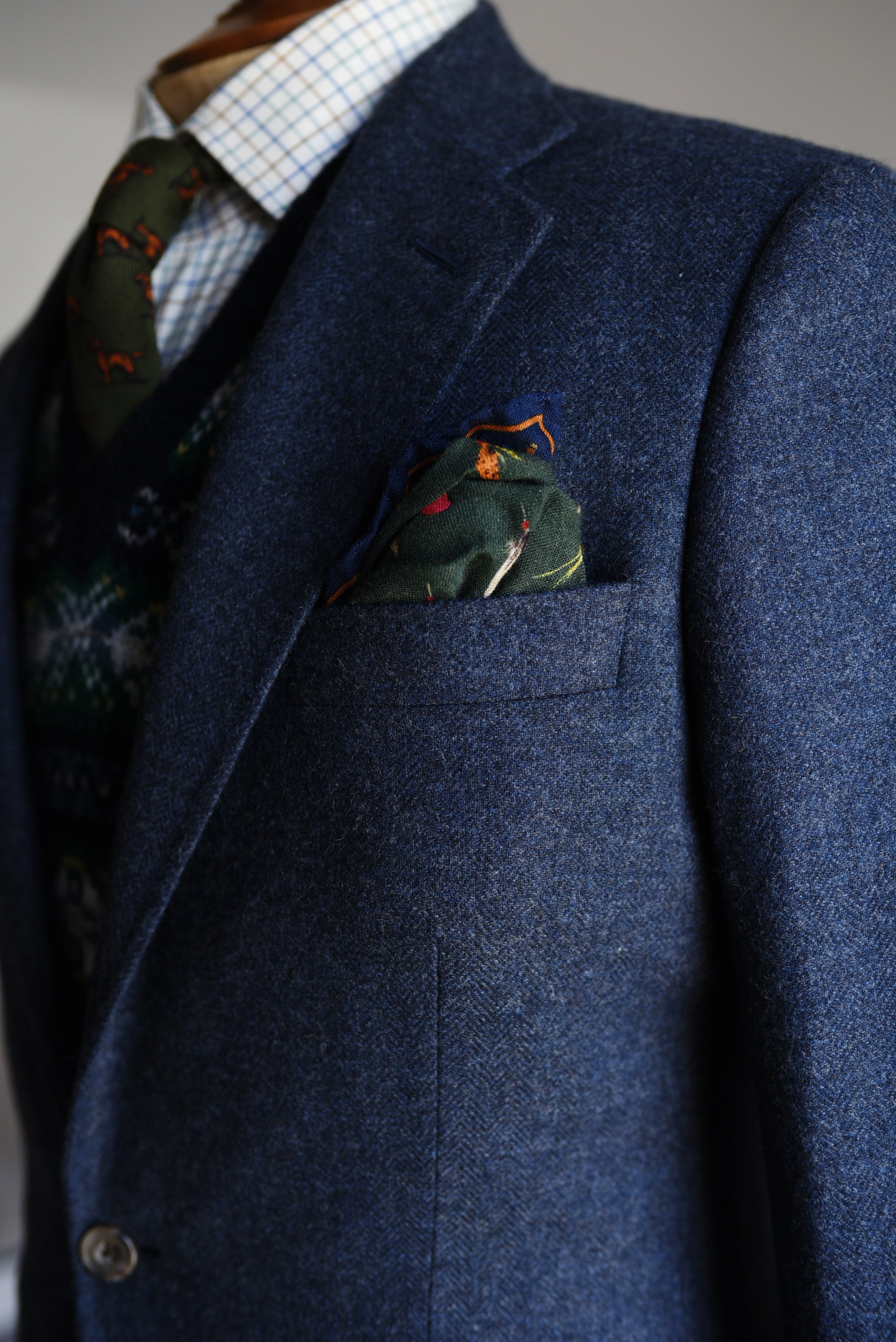 A close-up of a person wearing the Campbells of Beauly Navy House Jacket, 100% wool, styled with a patterned green pocket square, green tie with orange and brown designs, blue-and-white checked shirt, and a patterned sweater vest.