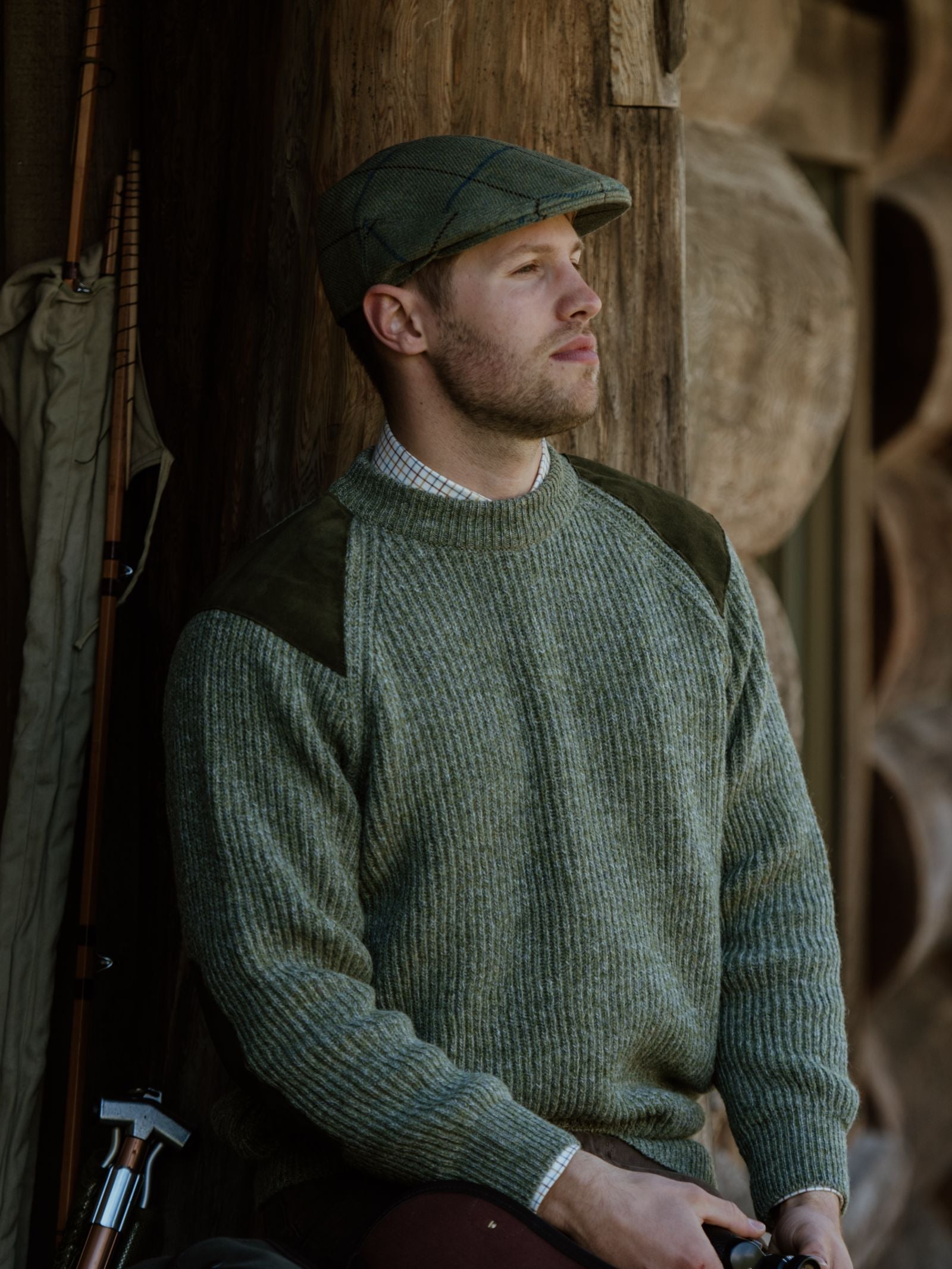 A man in a green flat cap and a Campbell’s of Beauly Scotia Suede Jumper with suede shoulder patches sits outdoors by a rustic wooden wall, gazing thoughtfully into the distance beside his fishing rod and gear.