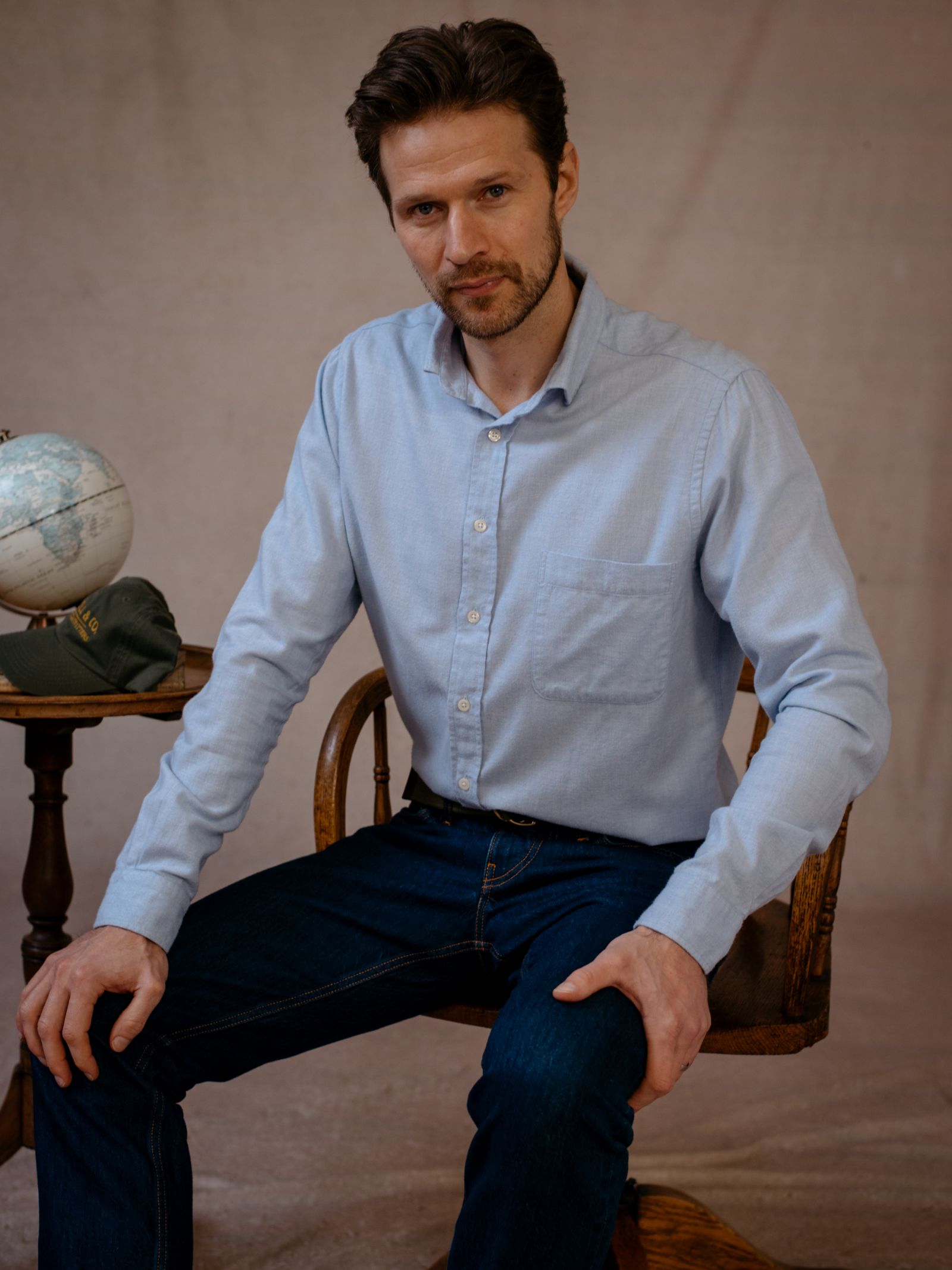 A man with short brown hair and a beard sits on a wooden chair, wearing the Campbells of Beauly Twill Shirt in light blue and dark jeans. His left arm rests on his knee; his right hand touches a table with a globe and green book. Beige background.