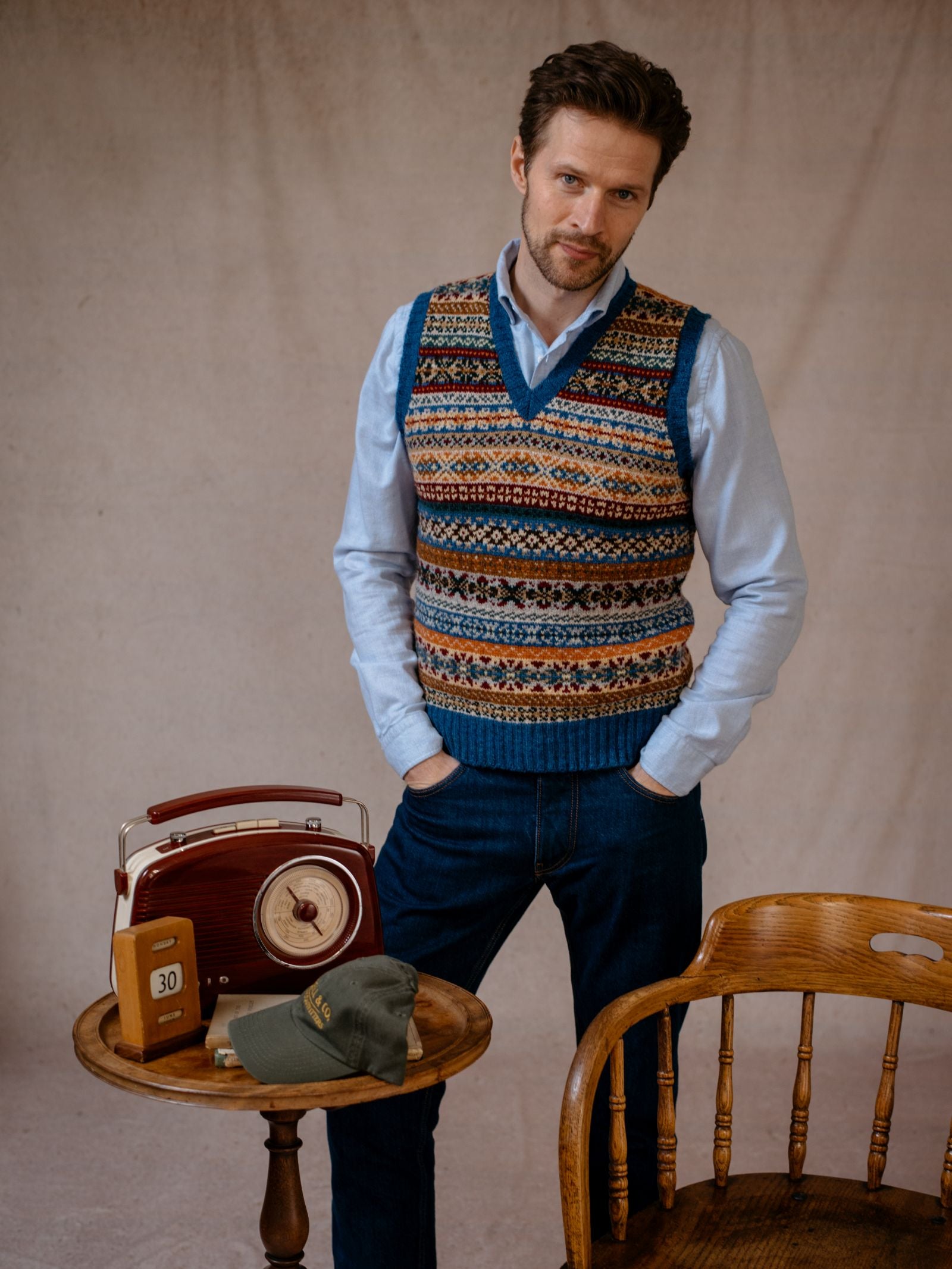 A man with short brown hair and a beard stands indoors wearing the Campbells of Beauly Fairisle Slipover over a blue shirt and jeans. He rests his hand on a table with a vintage radio, black book, and green cap; a wooden chair sits nearby.