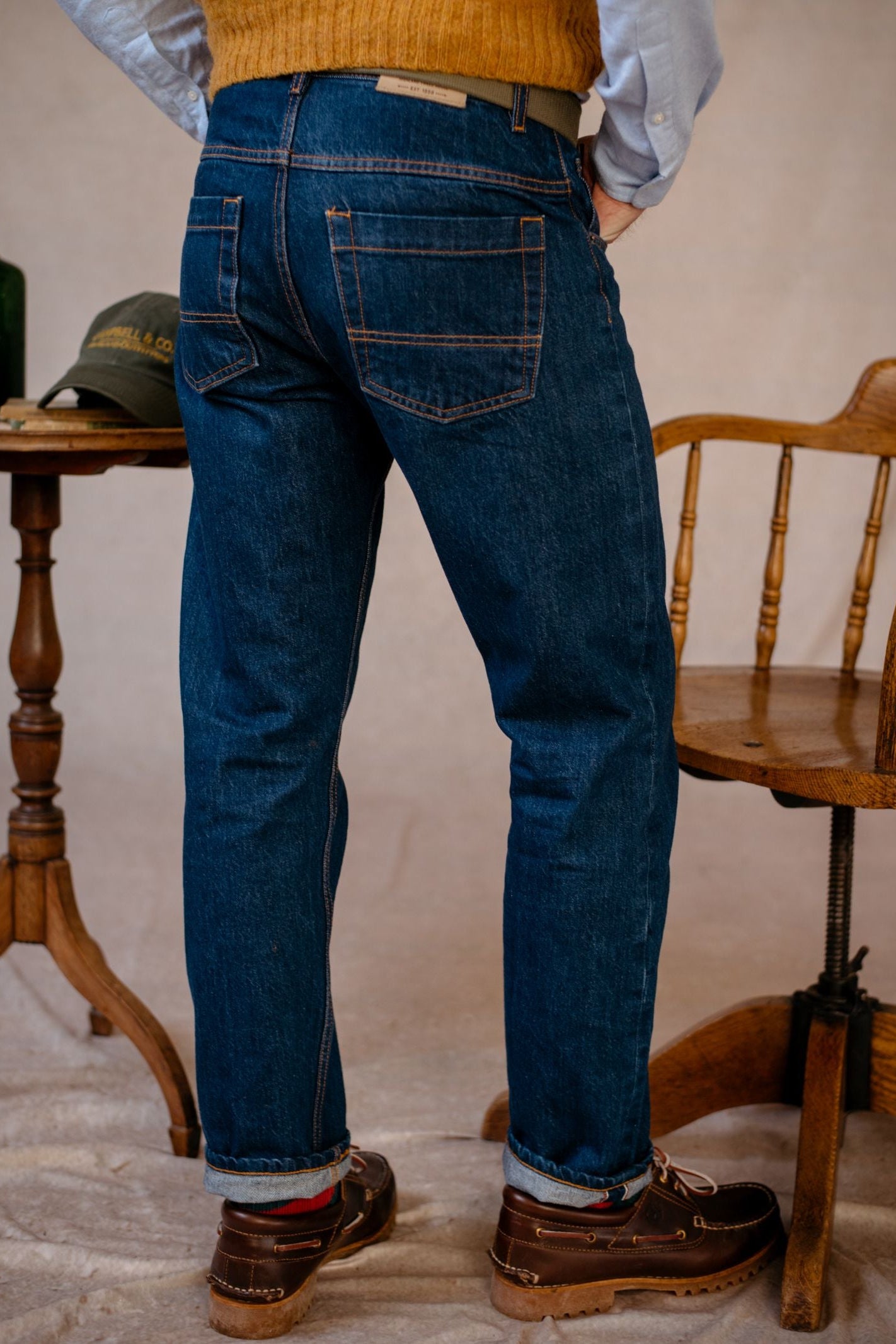 A person wearing Campbells of Beauly Denim Jeans, brown shoes, a light shirt, and a mustard sweater stands next to a wooden chair and a small round table with books and a bottle. The photo is taken from the back.