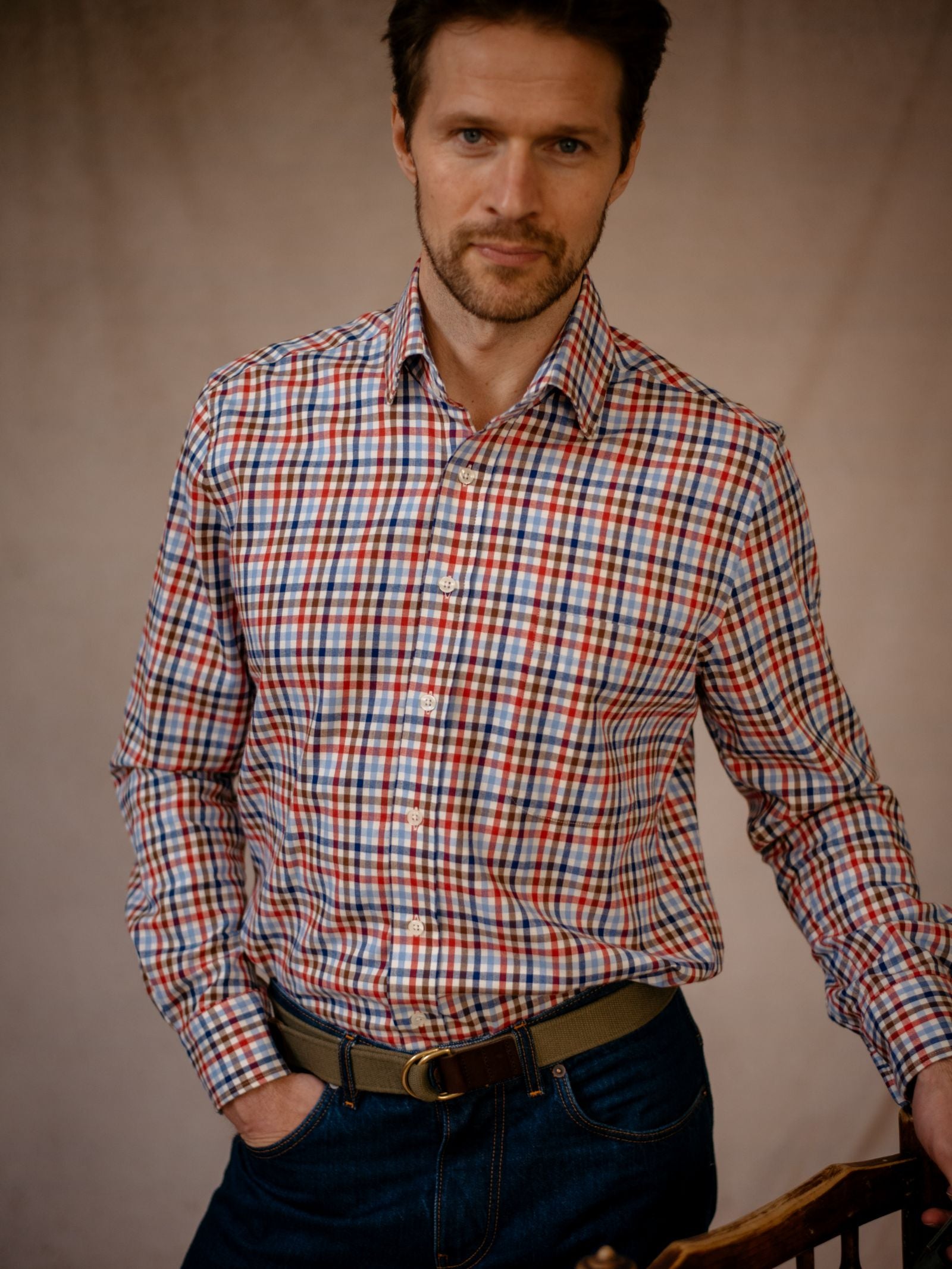 A man with short brown hair and a trimmed beard wears the Campbells of Beauly Windowpane Shirt—a red, blue, and white plaid brushed cotton shirt—tucked into dark jeans with a tan belt, standing casually against a neutral beige background.