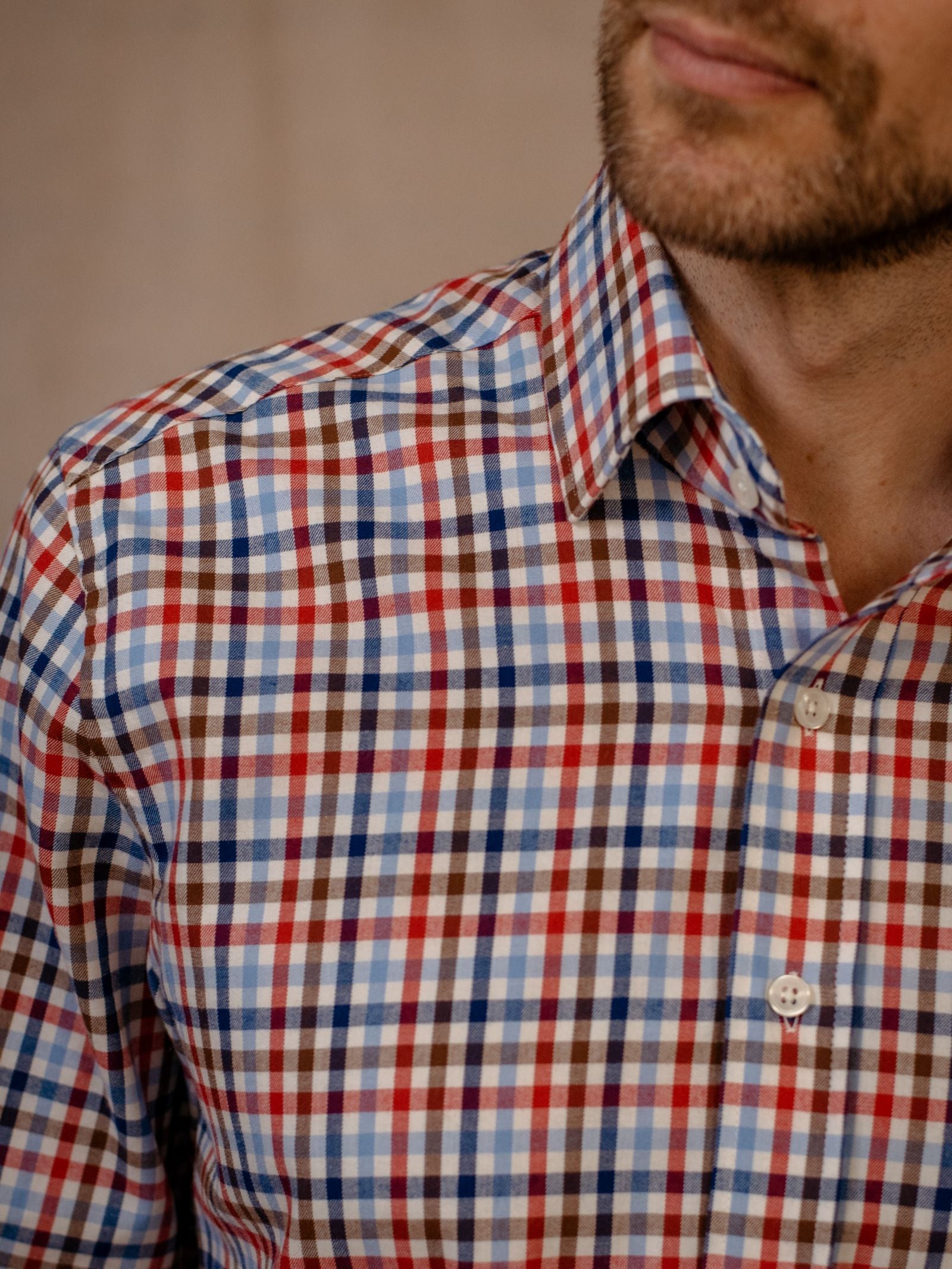 A close-up shows a man in the Campbells of Beauly Windowpane Shirt, a checkered button-up in red, blue, white, and brown brushed cotton. Only his lower face and upper torso are visible against a blurred neutral background.