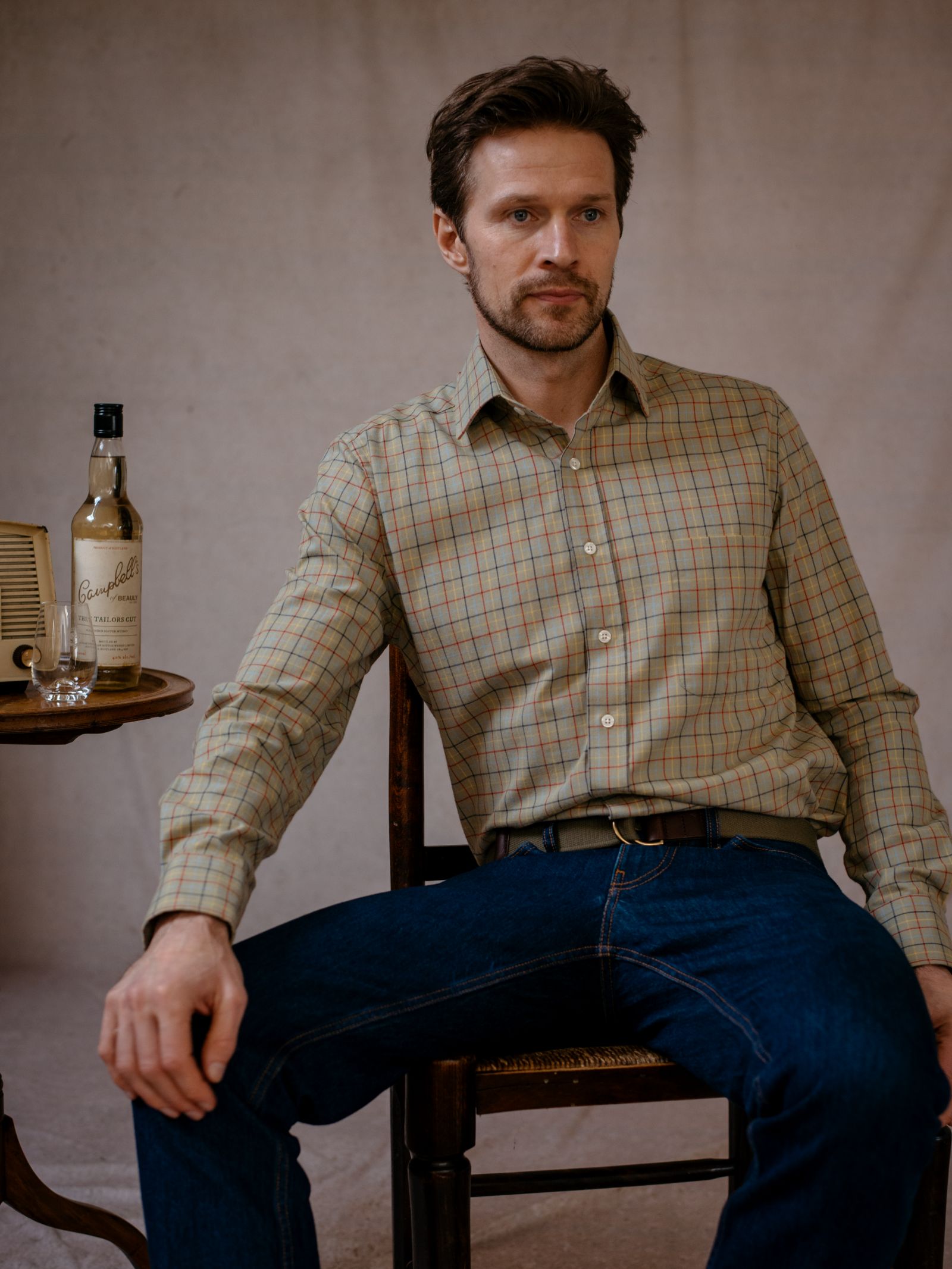 A man with short brown hair and a beard sits on a wooden chair, wearing the Campbells of Beauly Windowpane Shirt with dark blue jeans. A bottle of Scotch whisky and a glass are on a small table beside him. The background is plain and neutral.