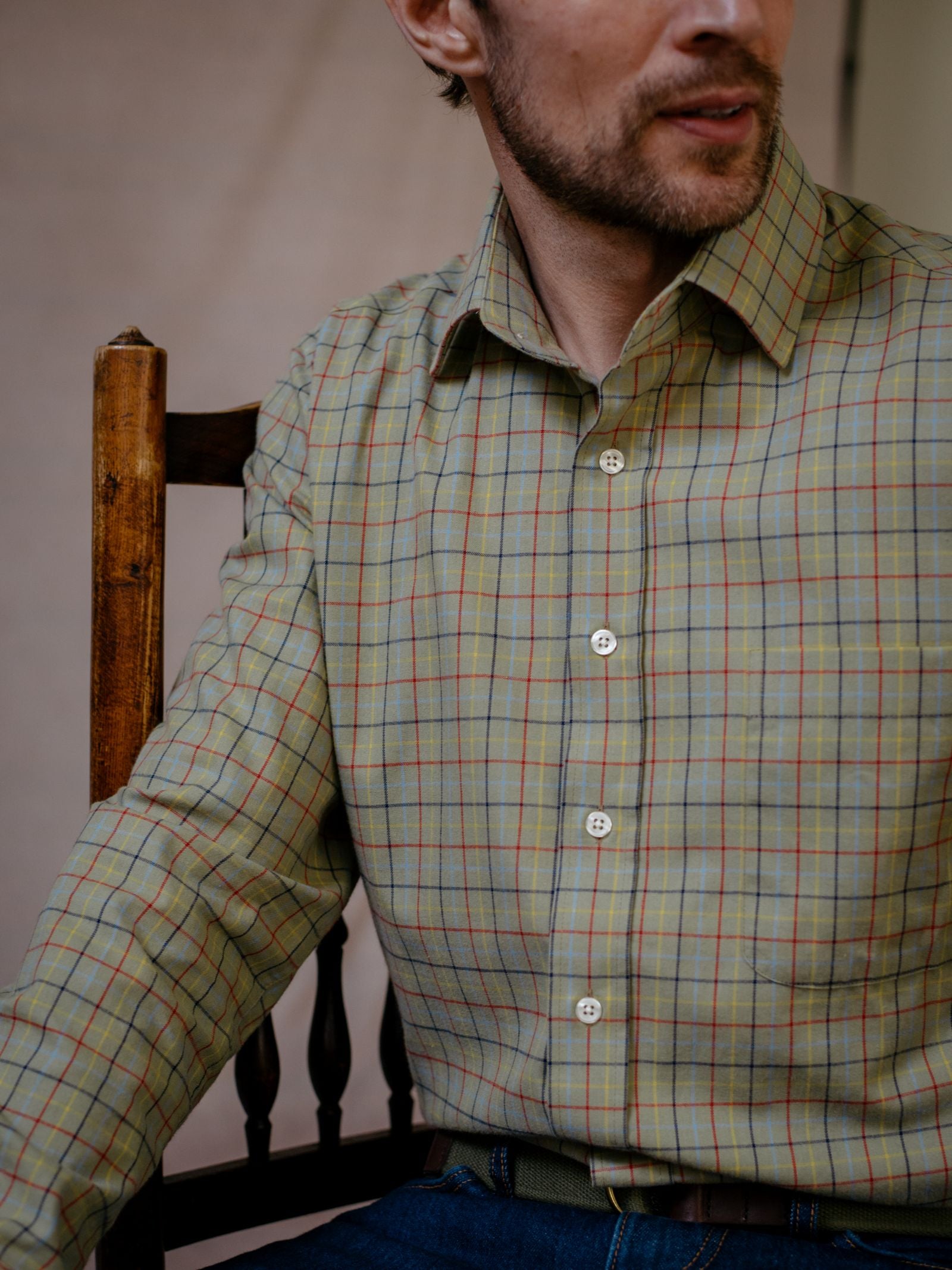 A man with light skin and short facial hair sits on a wooden chair, wearing the Campbells of Beauly Windowpane Shirt in green brushed cotton and blue jeans. Only the lower half of his face and upper torso are visible.