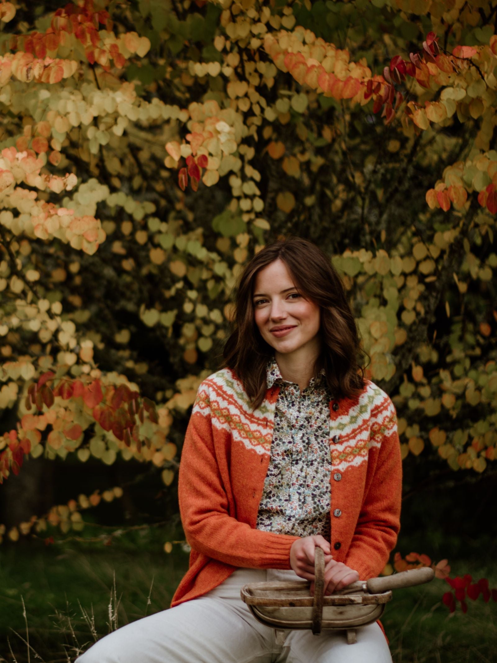 A woman with shoulder-length brown hair sits outdoors before autumn leaves, smiling gently as she wears a patterned orange Shetland Fairisle Cardigan by Campbell's of Beauly over a floral shirt and holds a rustic wooden basket.