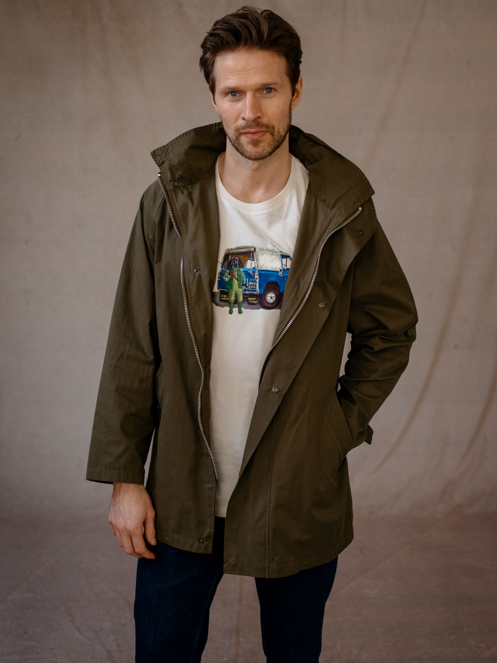 A man with short brown hair and a beard stands against a neutral backdrop, wearing the Mont Coat by Campbells of Beauly over a white T-shirt with a colorful blue vehicle graphic. He has one hand in his pocket and maintains a neutral expression.